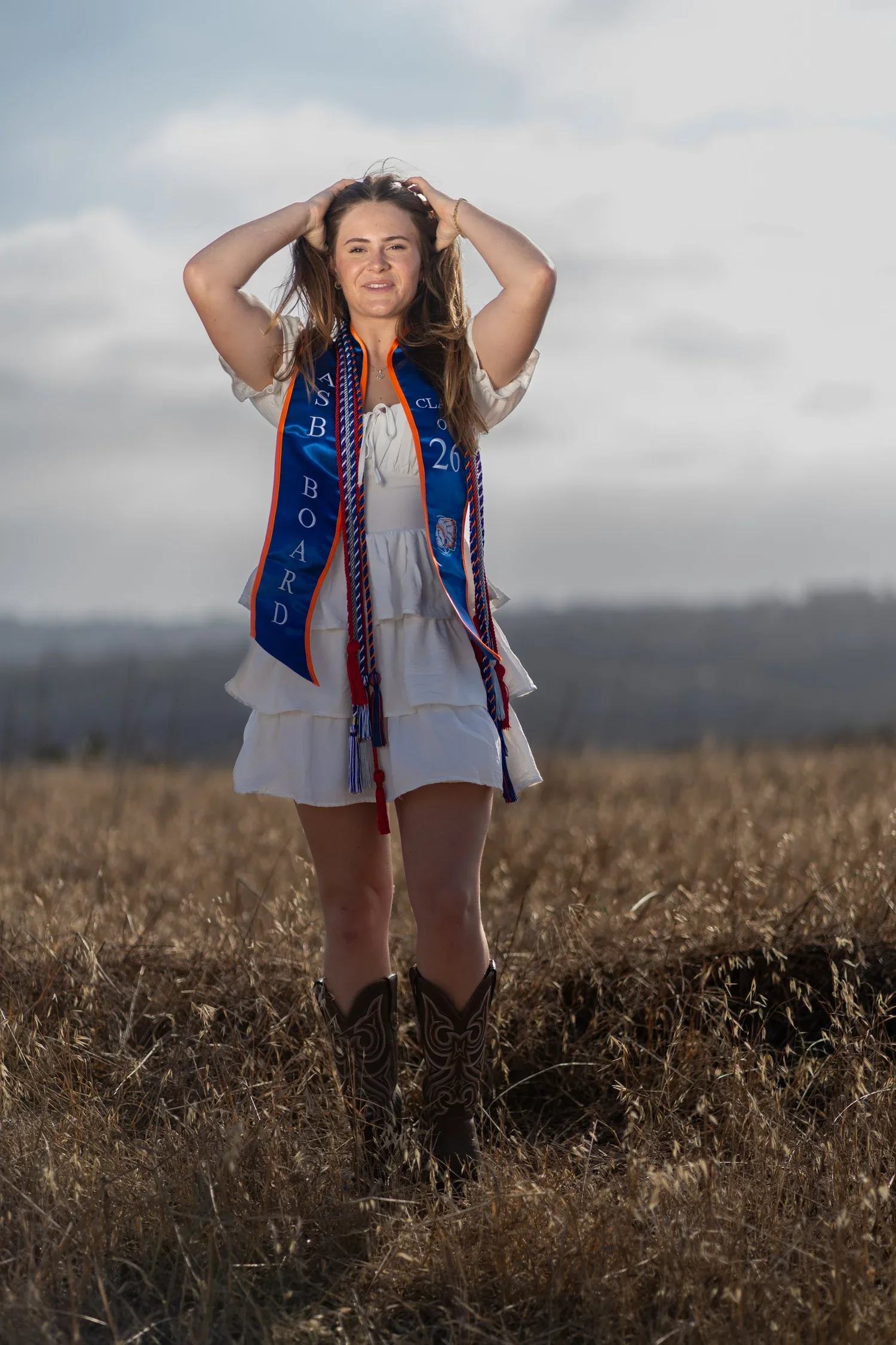 Young woman standing in a field of dry grass, wearing cowboy boots, a white dress, and a blue sash with orange trim and medals, raising her arms to hold her hair, with a cloudy sky in the background.
