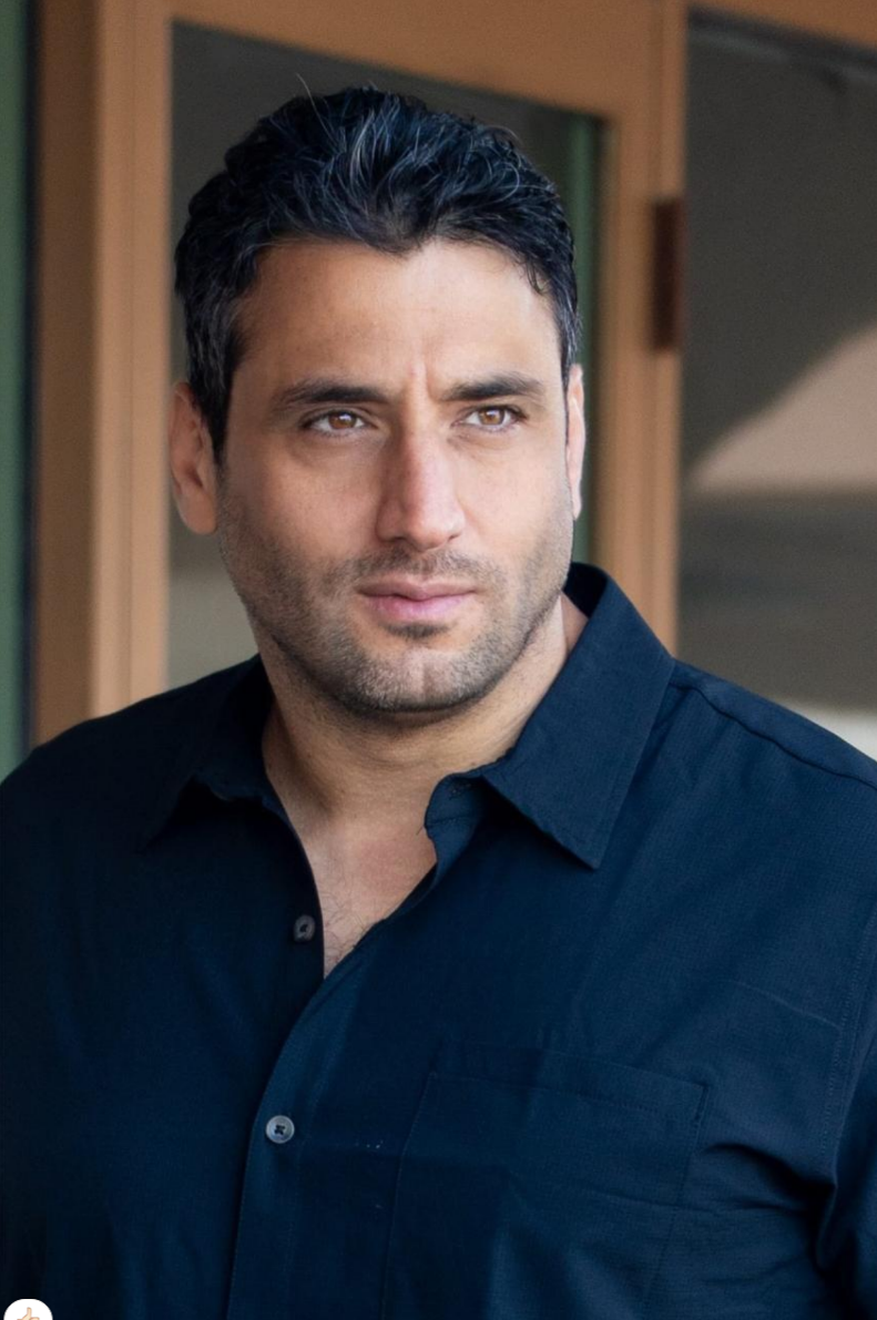 A man with dark hair and a beard, wearing a black collared shirt, looking to the side with a serious expression, standing outside a building.