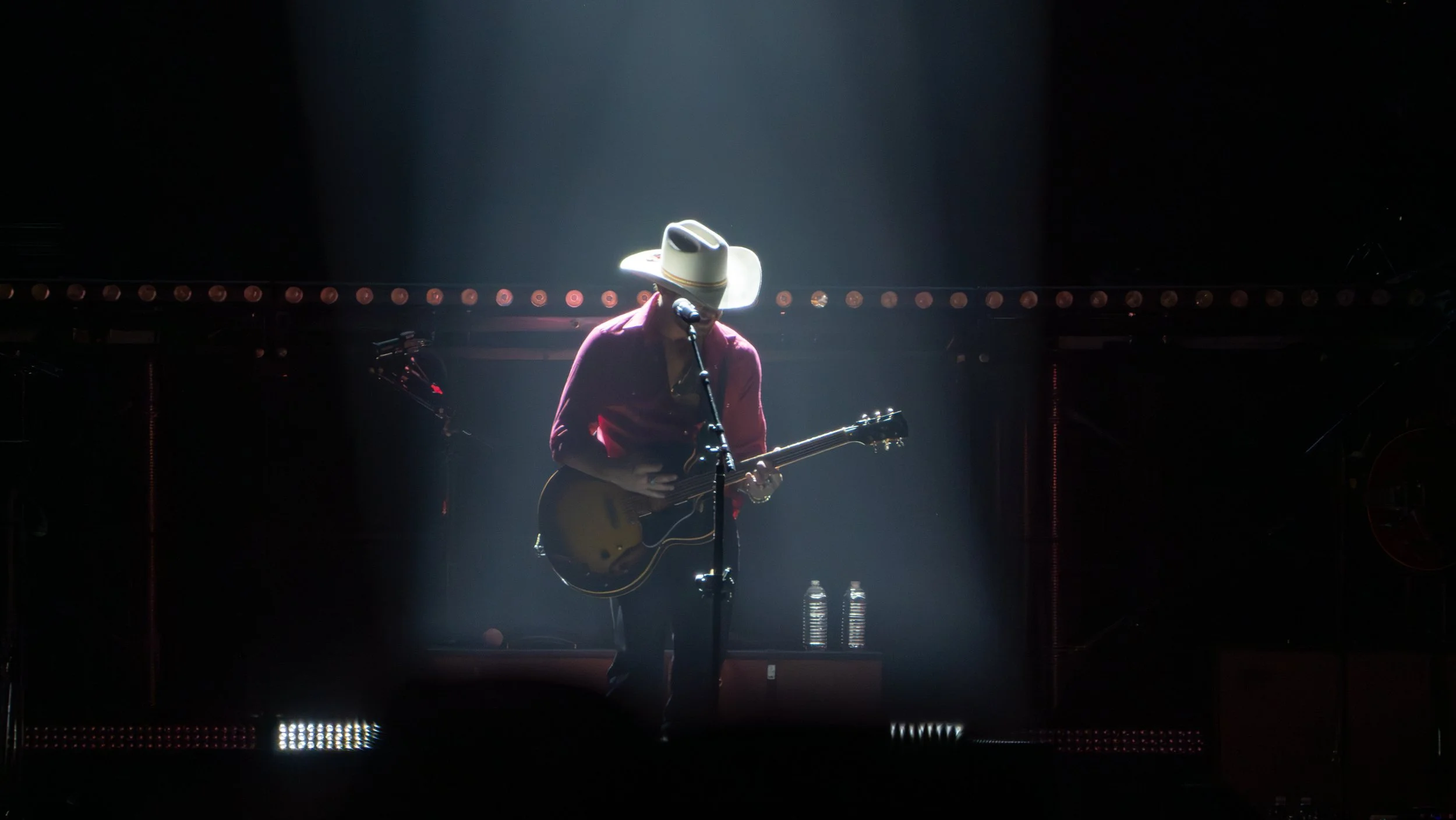 Performer in a cowboy hat playing guitar on stage with spotlight shining down, two water bottles on stage in front.