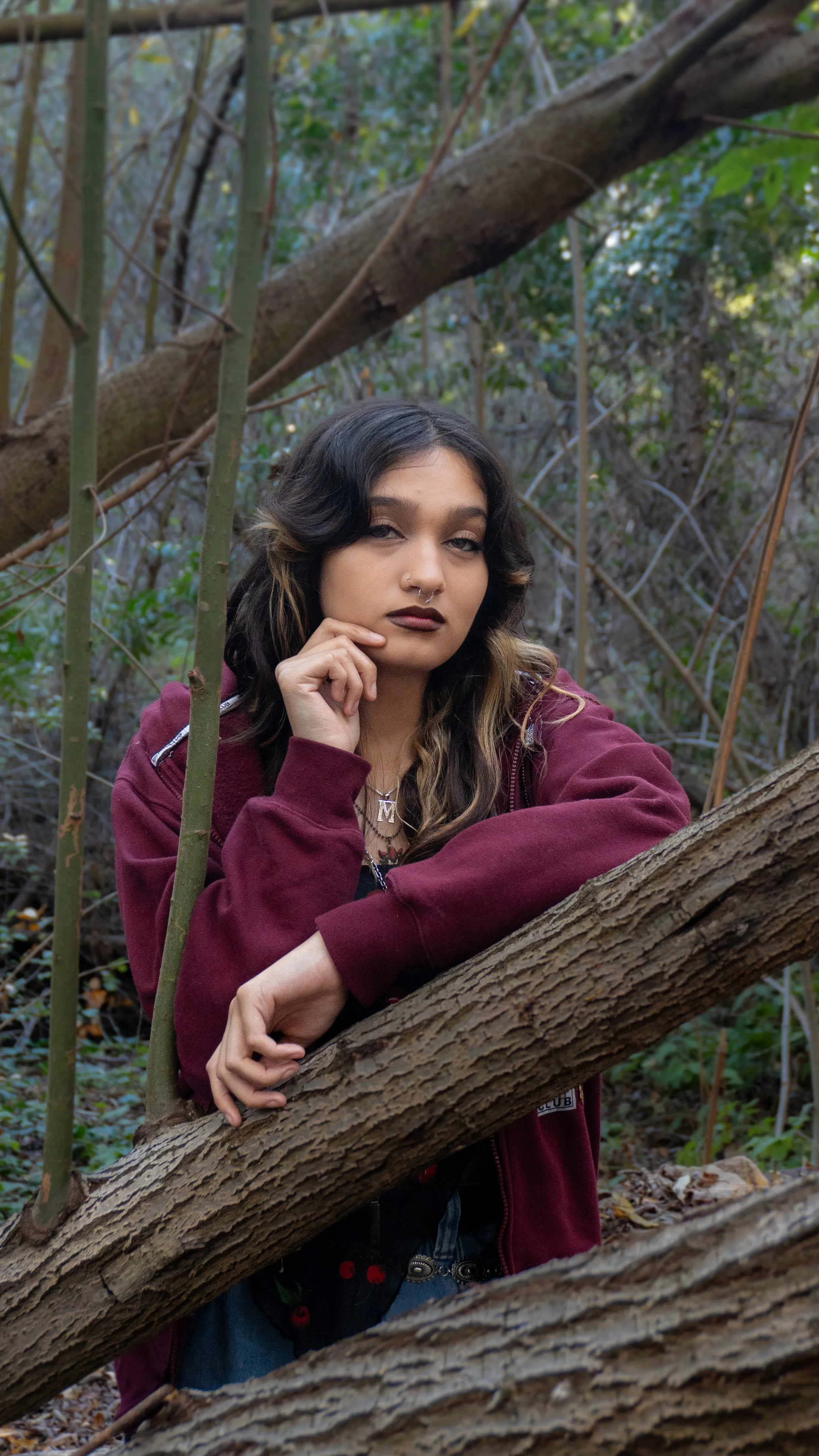 A young woman with dark hair, wearing a maroon jacket, sitting on a tree branch in a wooded area.