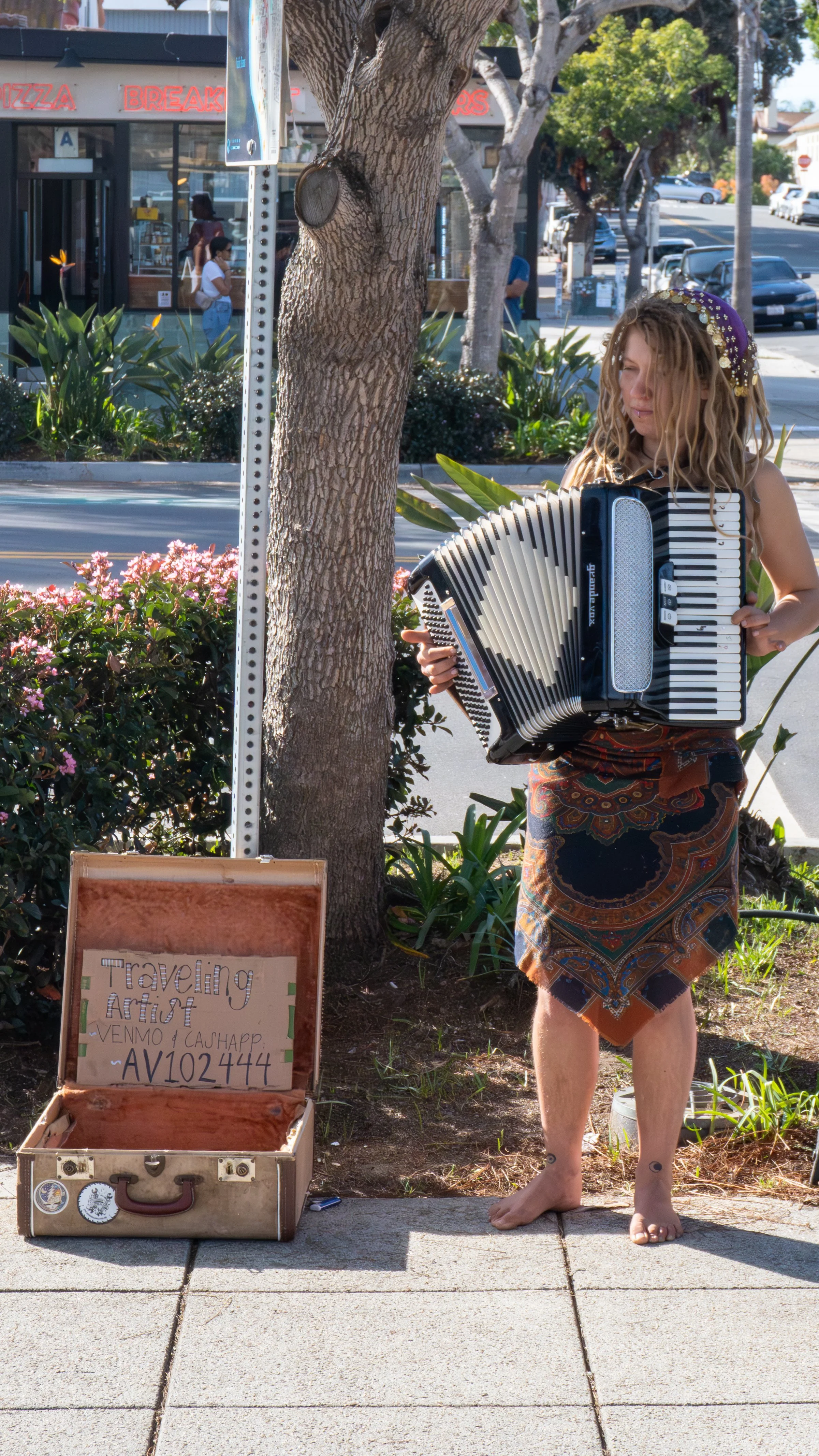 A barefoot woman with long, curly hair playing an accordion on a sidewalk street, with a sign in a suitcase reading 'Traveling Artist' and her Venmo and Cash App info. She is standing near a tree with a background of blurred pedestrians, parked cars, and a storefront.