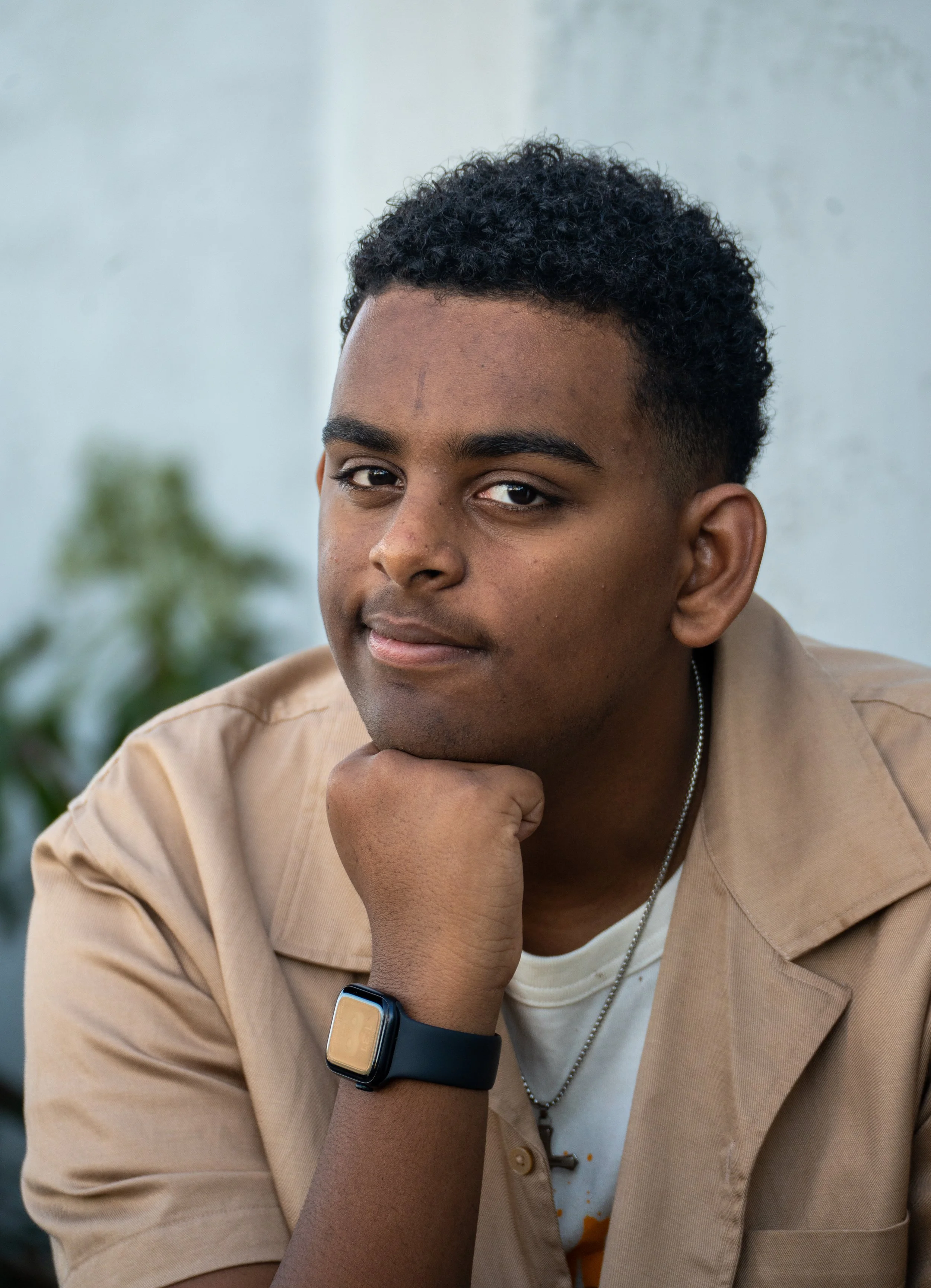 A young man with dark curly hair and medium skin tone, resting his chin on his hand, wearing a beige shirt and a smartwatch, looking at the camera with a confident expression.