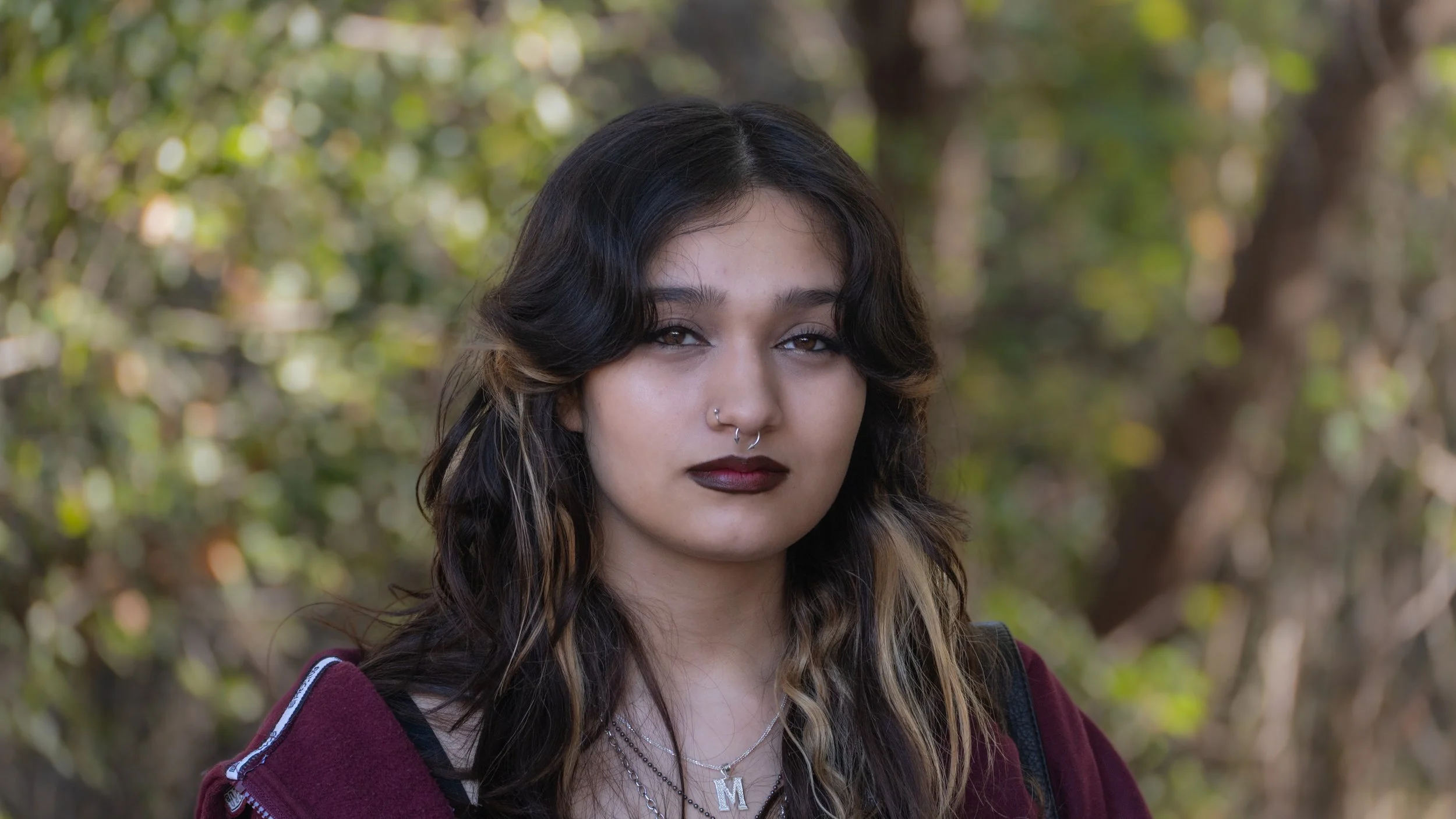 Portrait of a young woman with dark hair and brown highlights, wearing dark lipstick, silver jewelry, and a burgundy hoodie, standing outdoors with a background of trees and foliage.