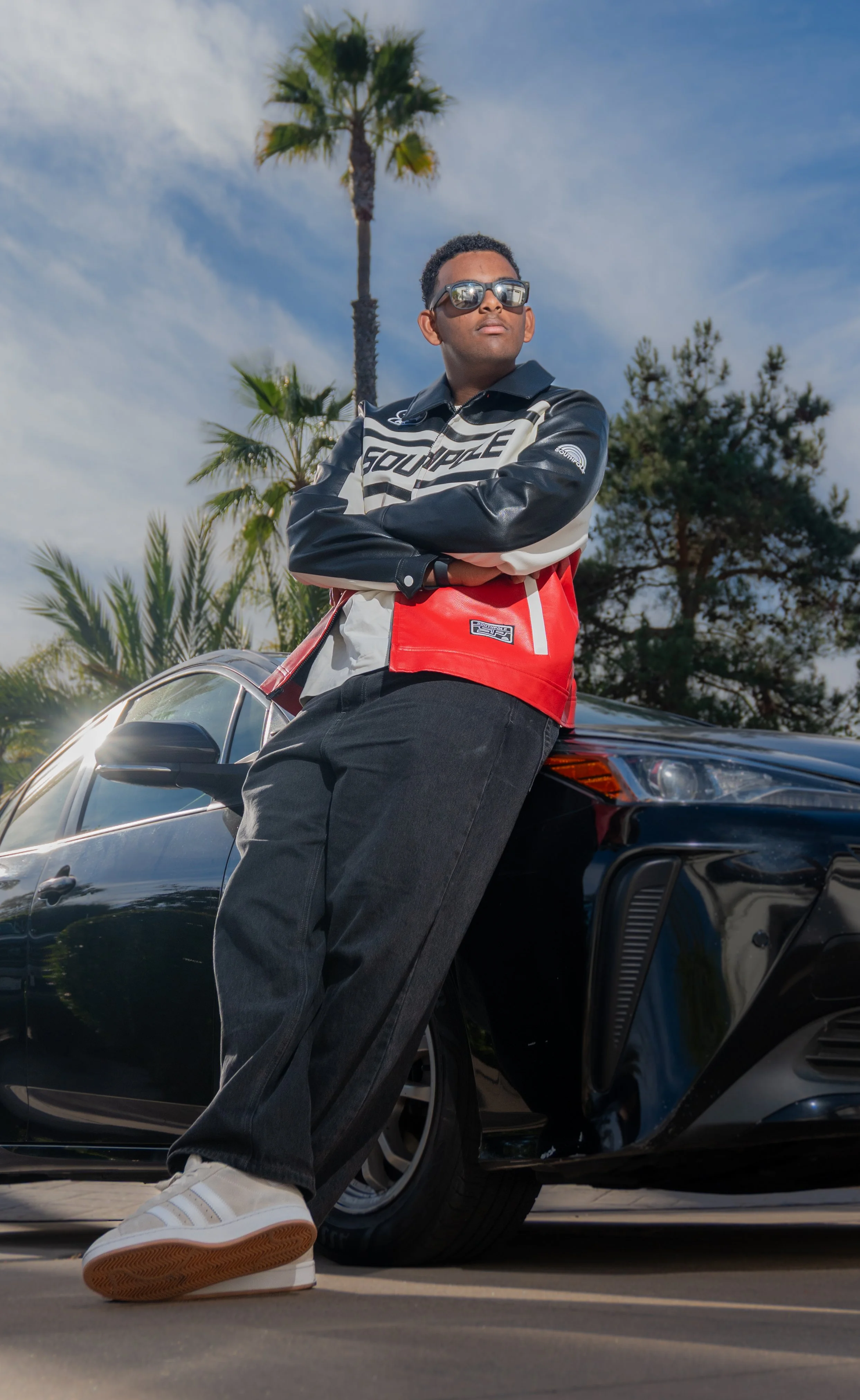 A young man wearing sunglasses, a black and white racing jacket, black jeans, and white sneakers leaning against a black sports car. Palm trees and a partly cloudy sky are in the background.