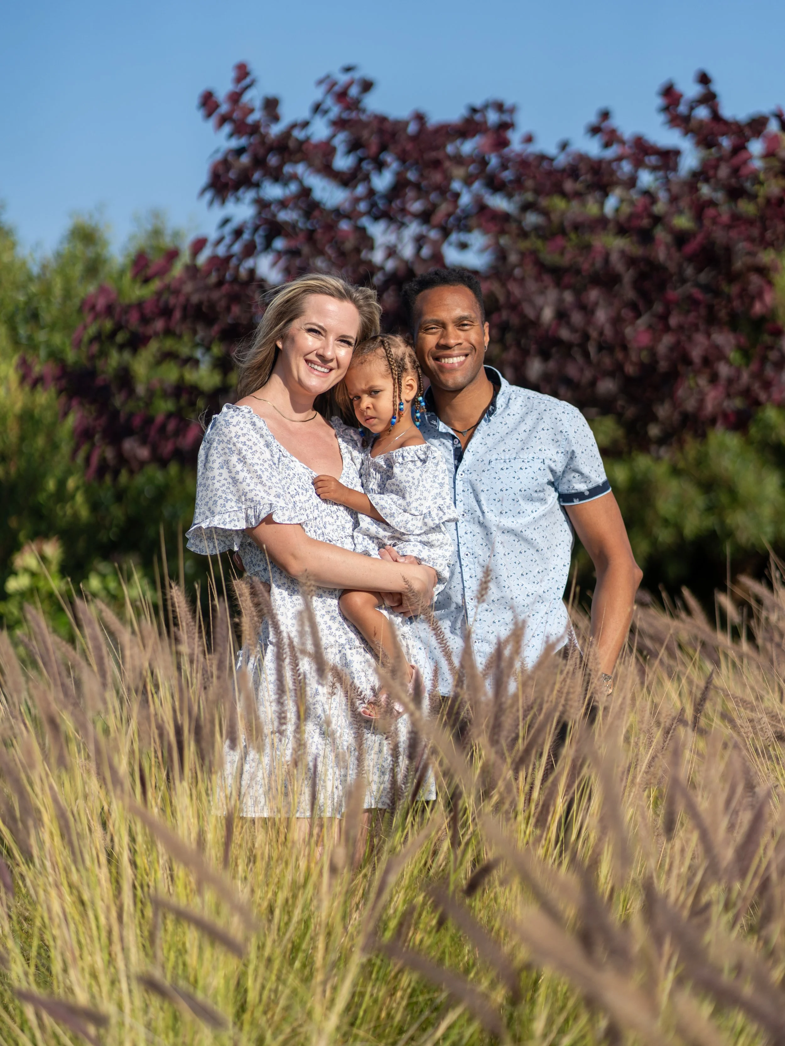 A family of three standing in a field of tall grass with trees and a blue sky in the background, smiling at the camera.