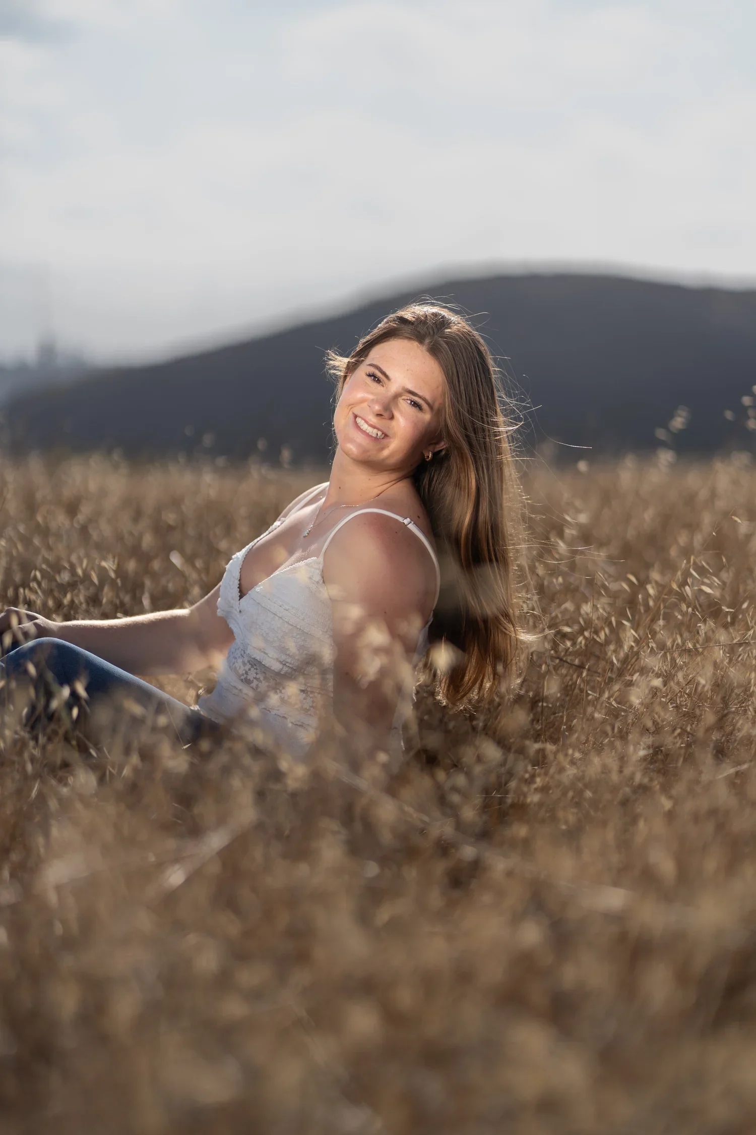 A young woman with long brown hair and a white top sitting in a field of dry grass, smiling at the camera with a mountain in the background.