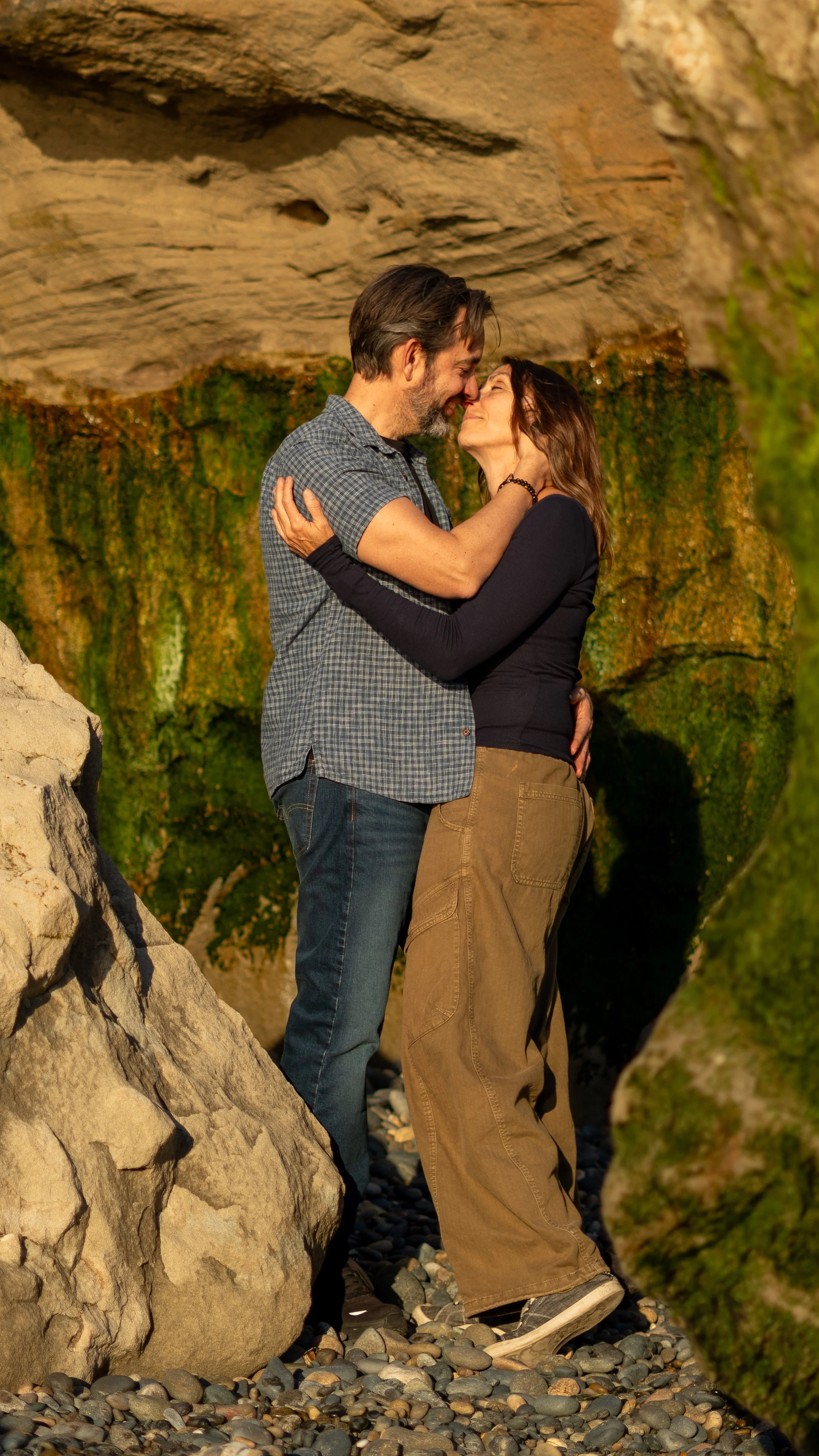 A couple embracing and about to kiss on a rocky beach with moss-covered rocks and cliffs in the background.