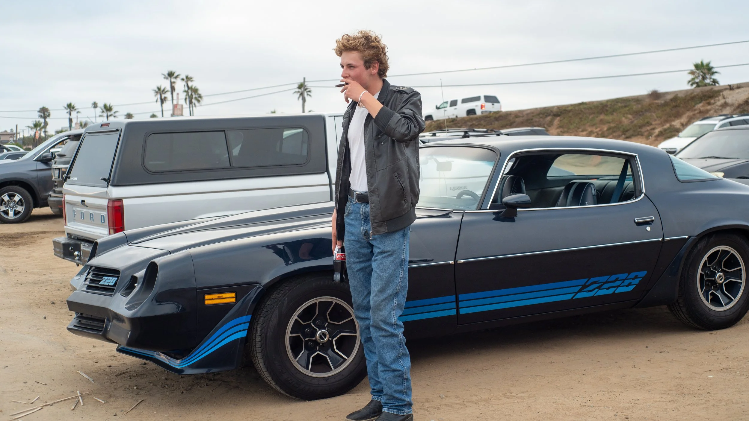 A man standing next to a vintage black car with blue racing stripes, smoking a cigarette, in a parking lot with other vehicles and palm trees in the background.