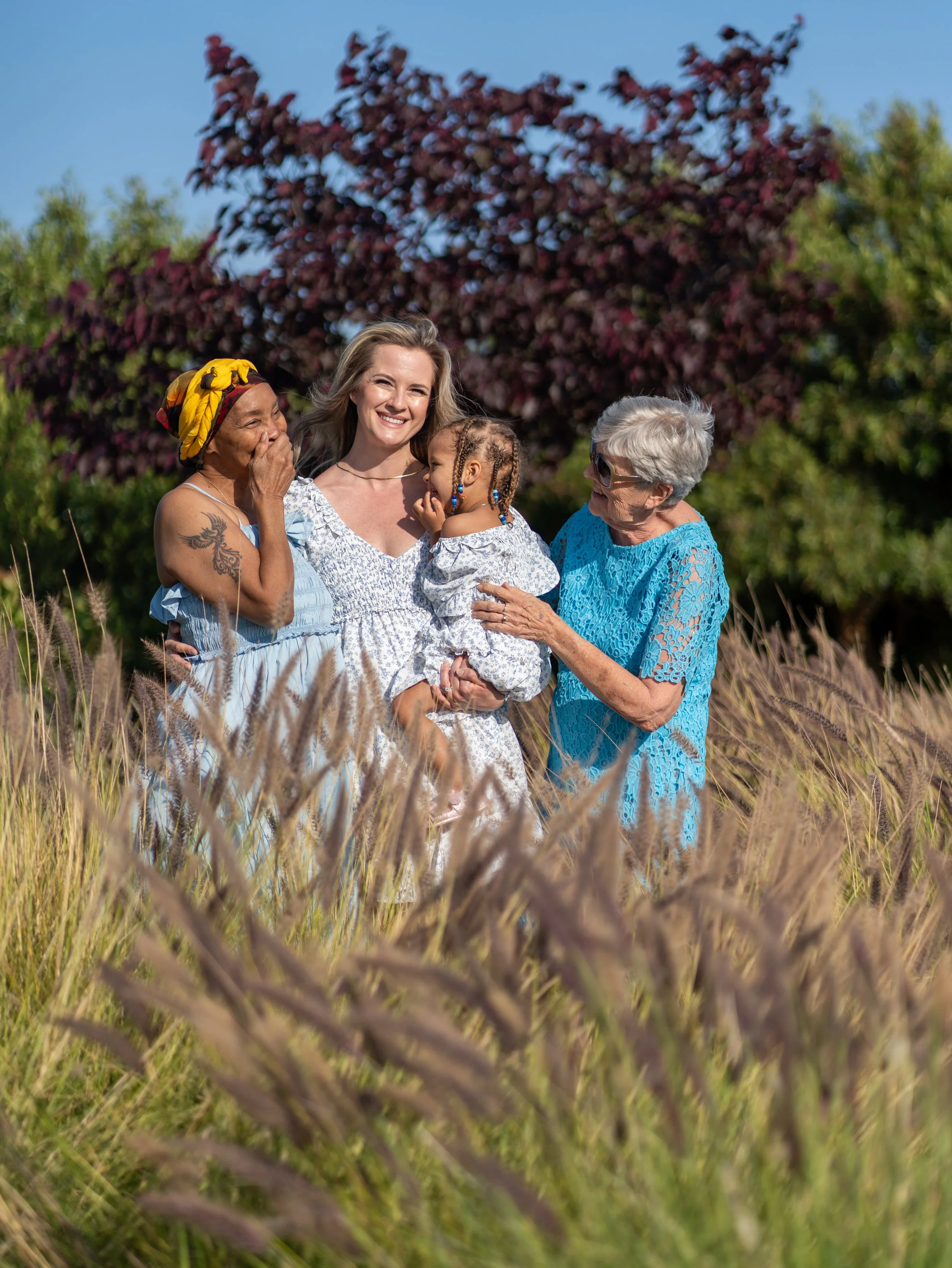 A group of women and a young girl smiling and laughing together outdoors in a field with tall grass, with trees and a bright blue sky in the background.
