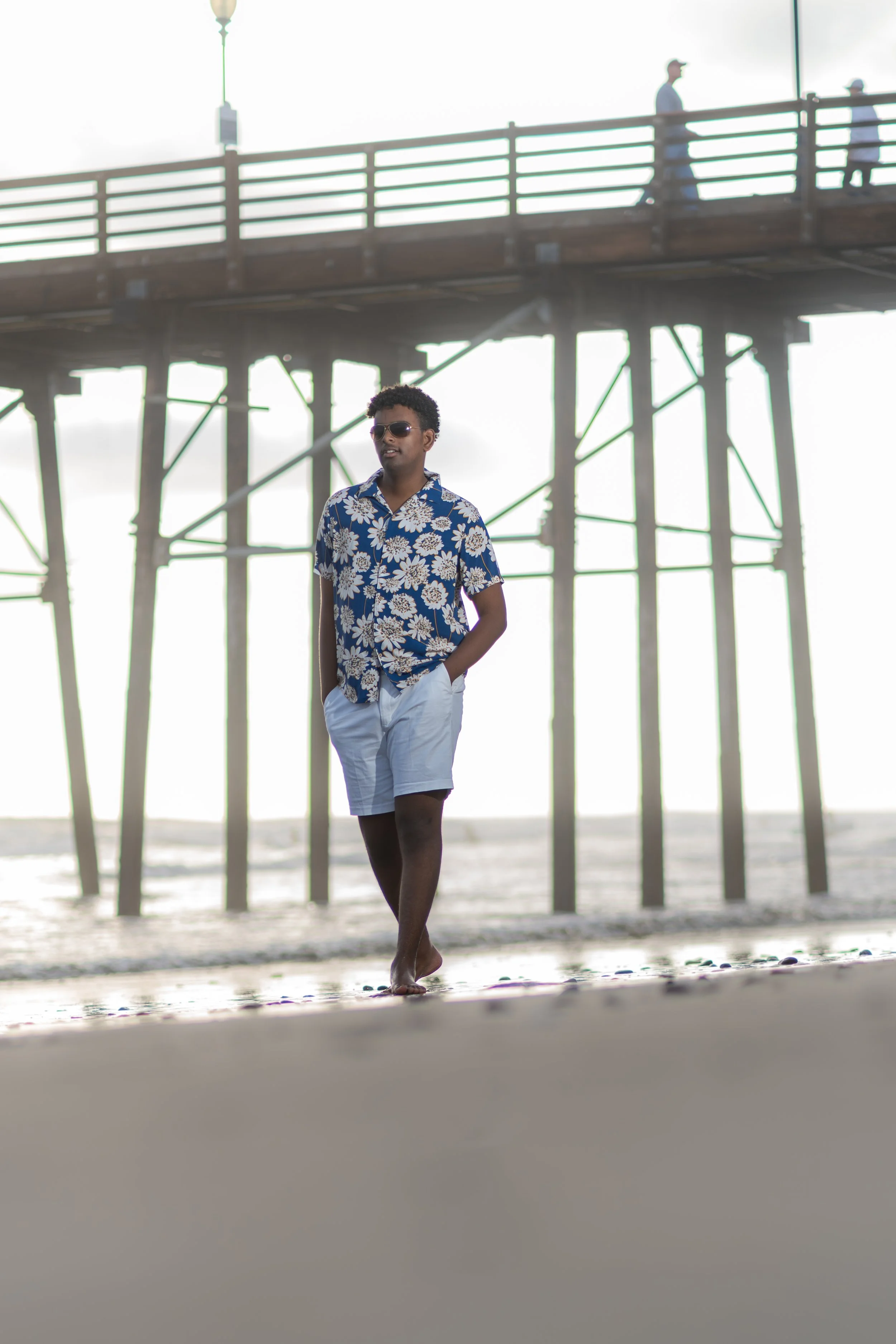 Young man walking barefoot on the beach near a pier, wearing sunglasses, a blue Hawaiian shirt, and white shorts, with a bright sky and ocean in the background.