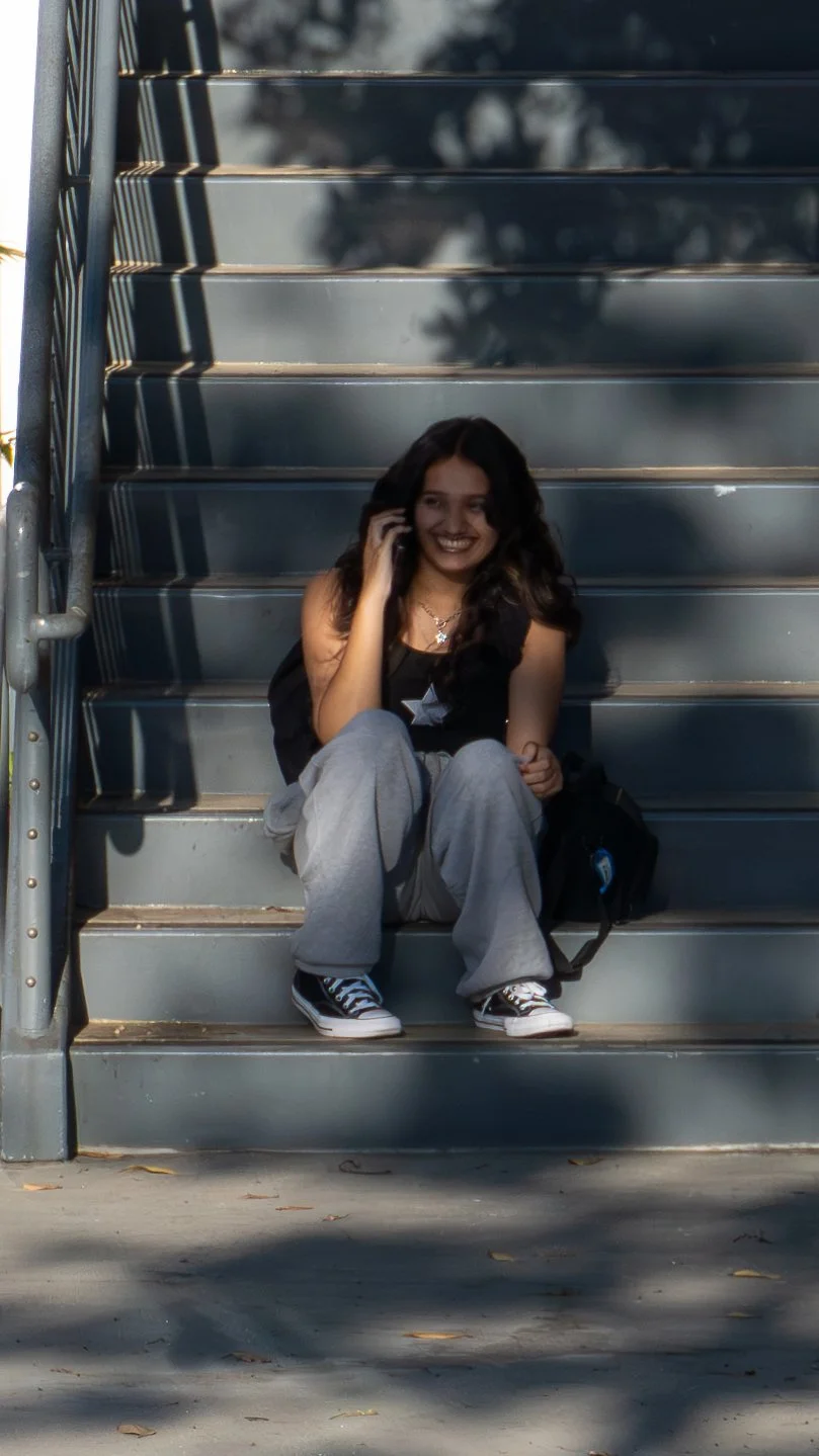 A young woman sitting on outdoor stairs, talking on her phone and smiling, with a backpack beside her.