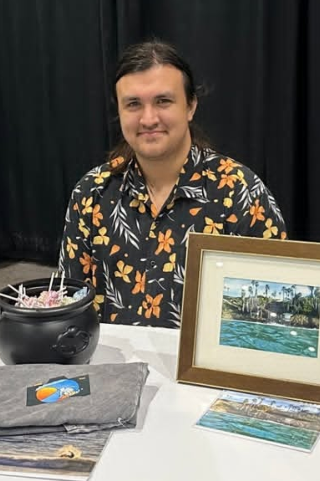 A man with long dark hair wearing a black Hawaiian shirt with orange and white floral pattern sitting at a table with framed artwork and an ice cream pot.