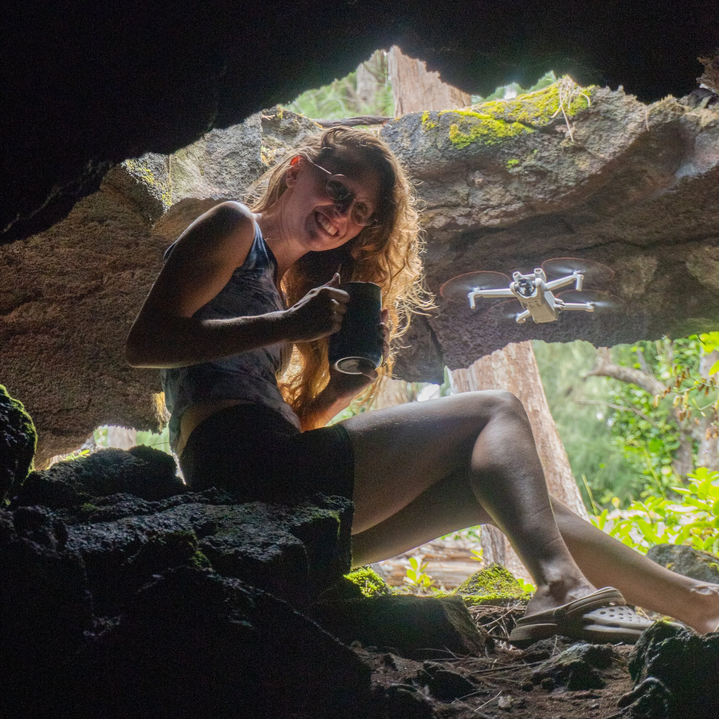 A woman with curly red hair and sunglasses sitting inside a cave, smiling and holding a black mug, with a drone flying nearby.