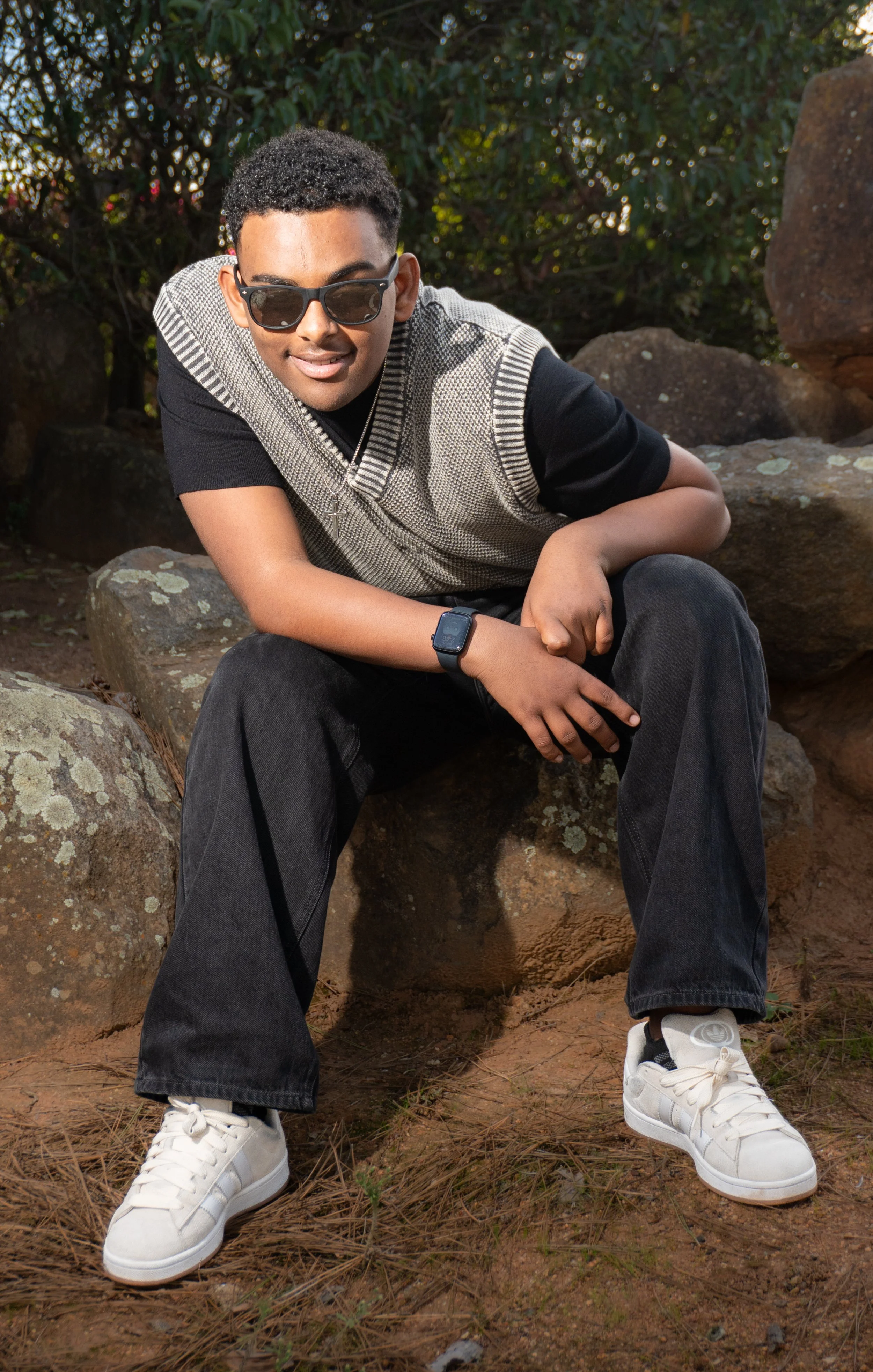 A young man with dark curly hair, wearing sunglasses, a black and gray vest, black pants, white sneakers, and a smartwatch, sitting on rocks outdoors with greenery in the background.