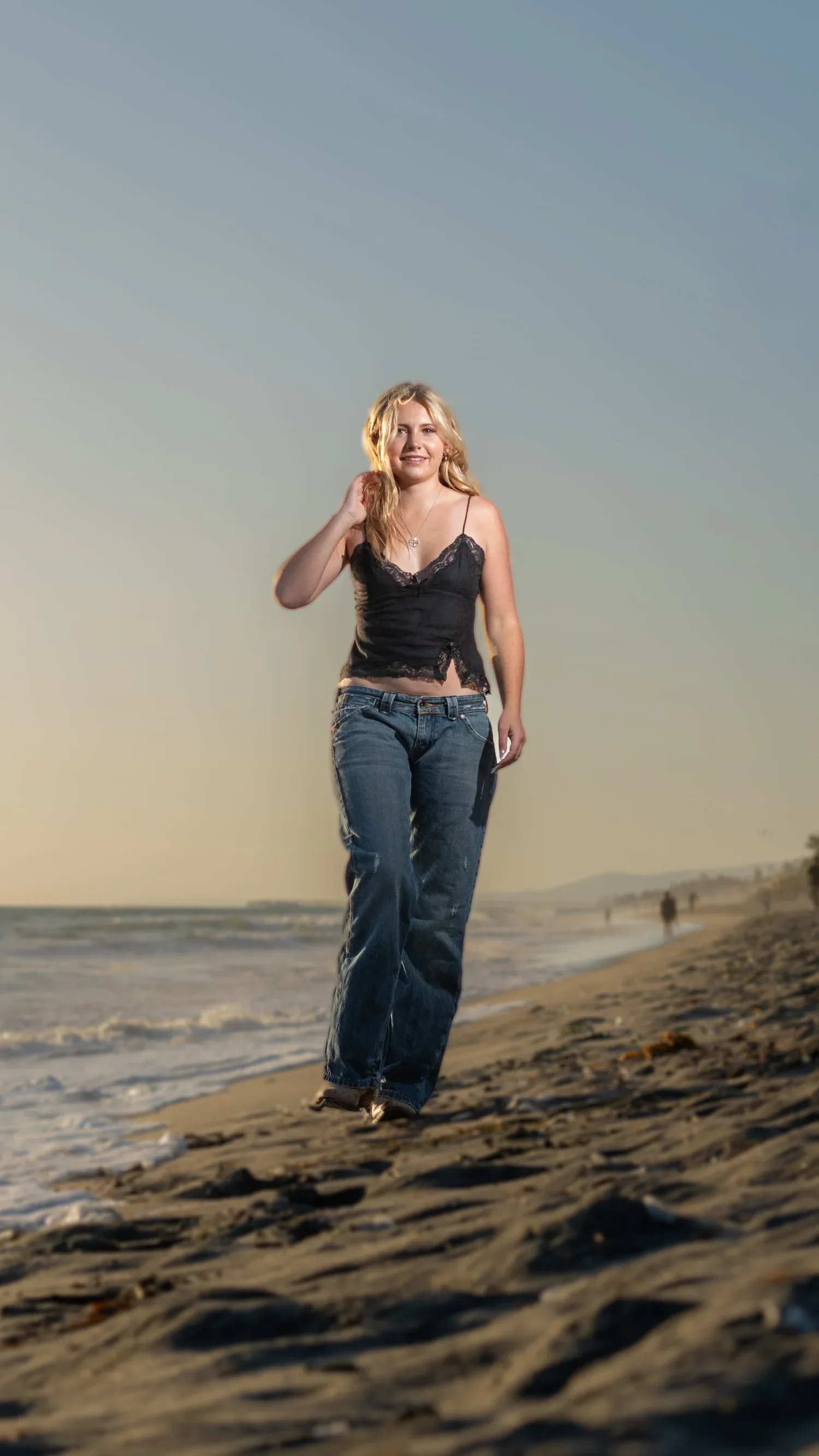 A young woman in a black lace top and jeans walking along a beach during sunset.