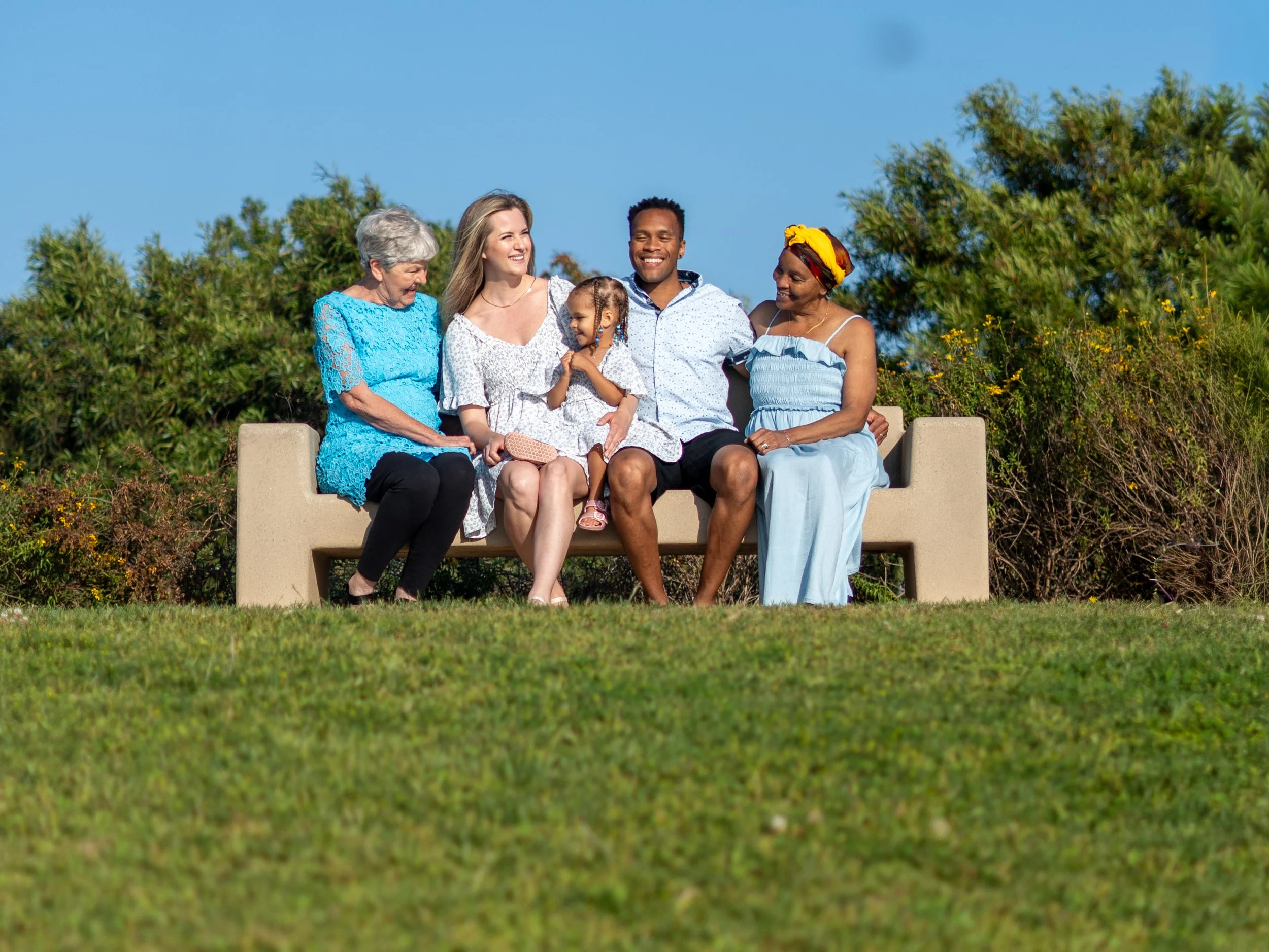 A group of five people, including elderly, adults, and a child, sitting on a bench outdoors against green shrubbery and a blue sky, smiling and enjoying each other's company.