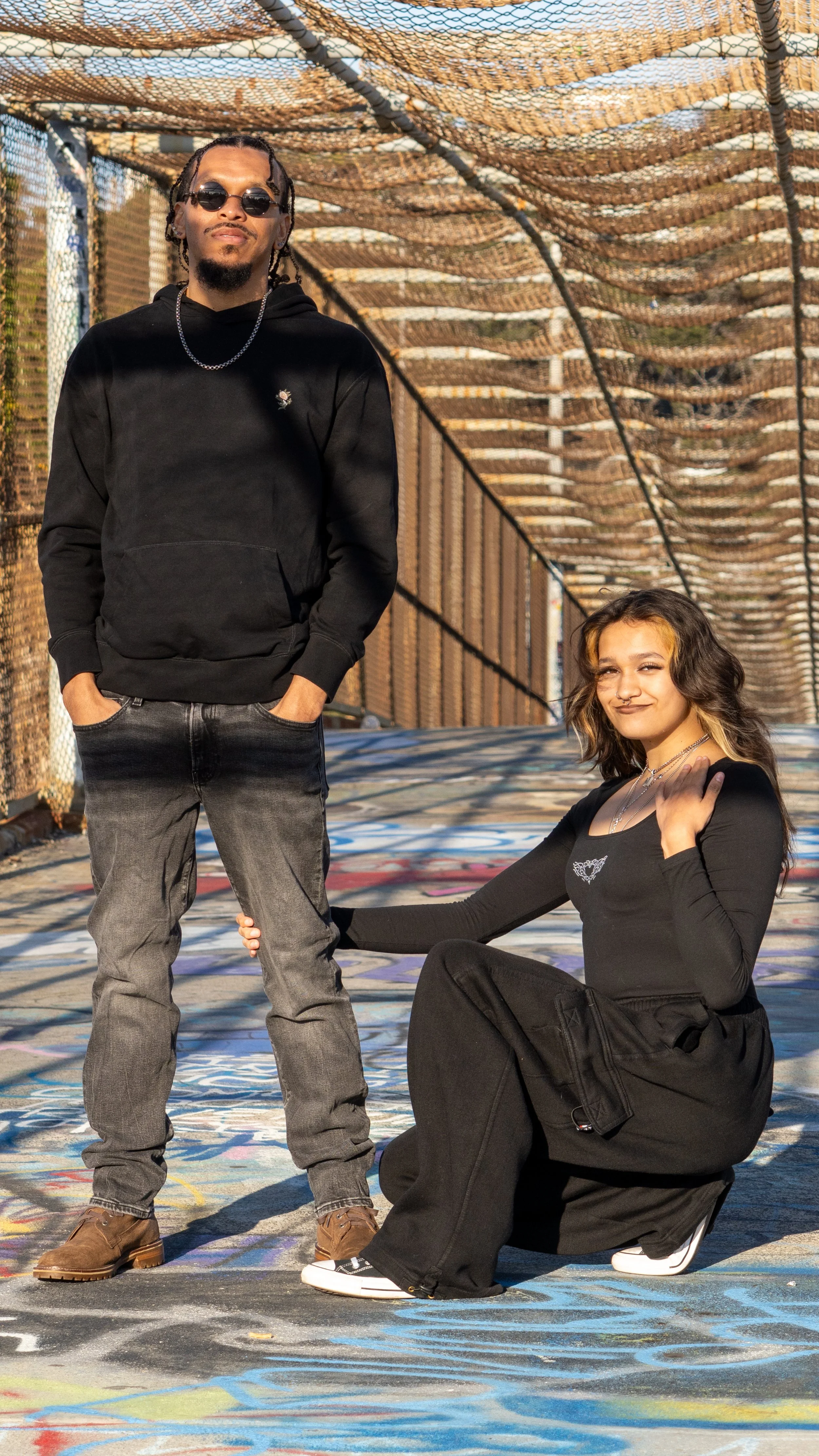 A young man and woman pose on a graffiti-covered outdoor bridge. The man stands with hands in pockets, wearing sunglasses, a black hoodie, gray jeans, and brown boots. The woman is sitting on the ground, with her hand on the man's leg, wearing a black long-sleeve shirt, black pants, and sneakers, with wavy hair and a confident smile.