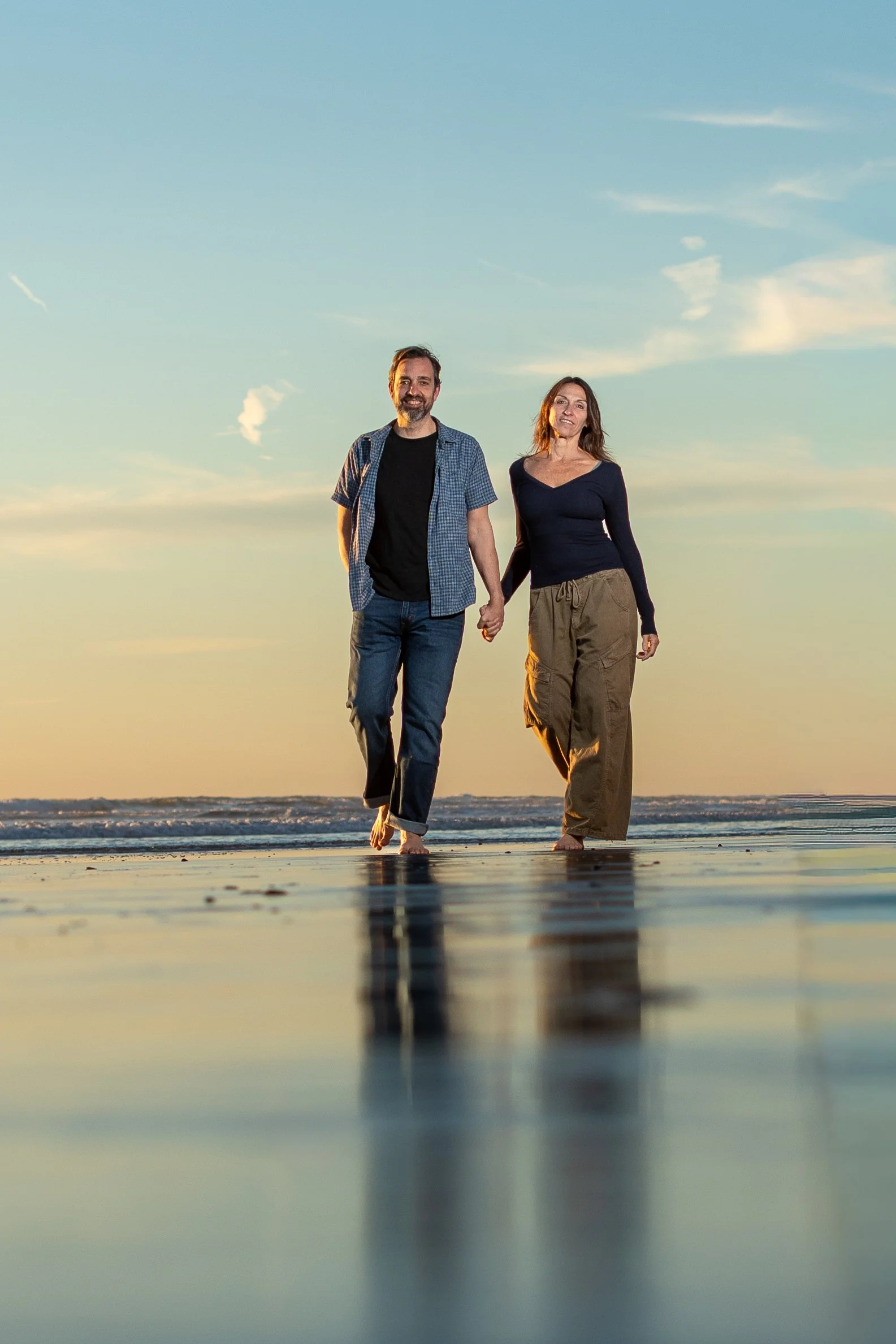 A couple walking hand in hand on the beach during sunset, barefoot, with the sky and ocean in the background.