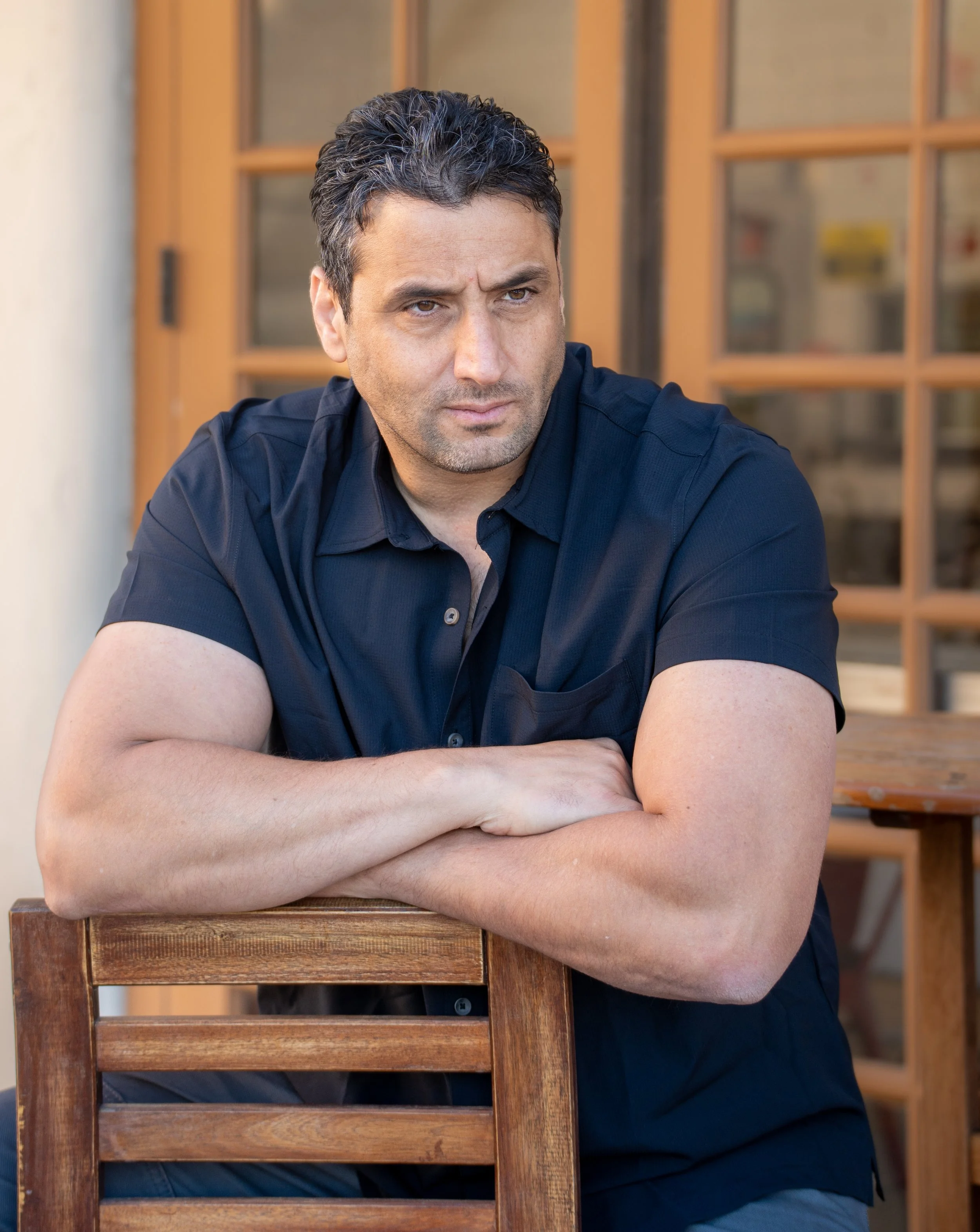 A man with dark hair and a beard sits outdoors at a wooden table, wearing a dark blue short-sleeved shirt, with arms crossed on the back of a wooden chair.