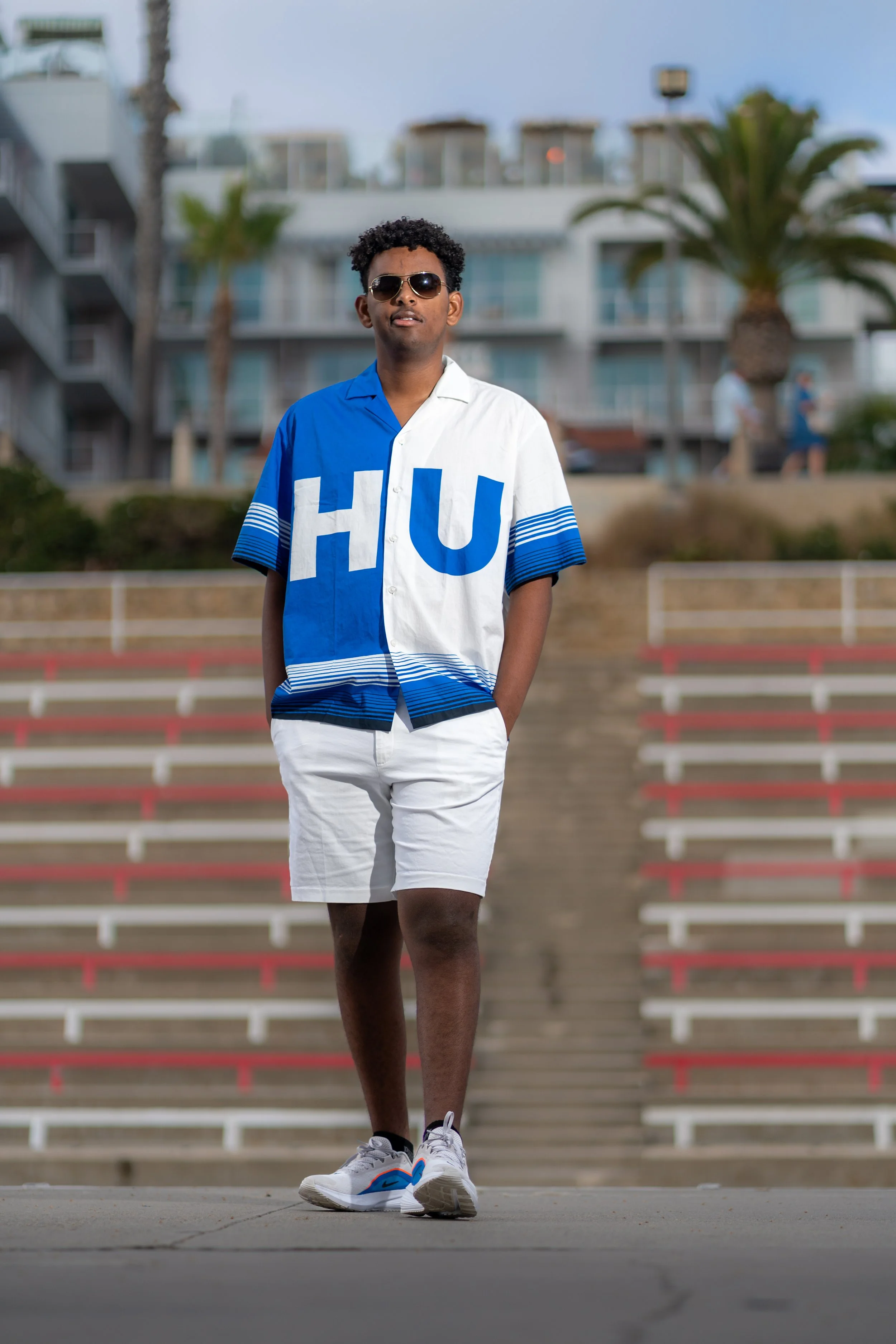 A young man with curly hair wearing sunglasses, a two-tone blue and white short-sleeve shirt with the letters 'HU' on it, white shorts, and sneakers, standing in an outdoor sports stadium with bleachers and palm trees in the background.