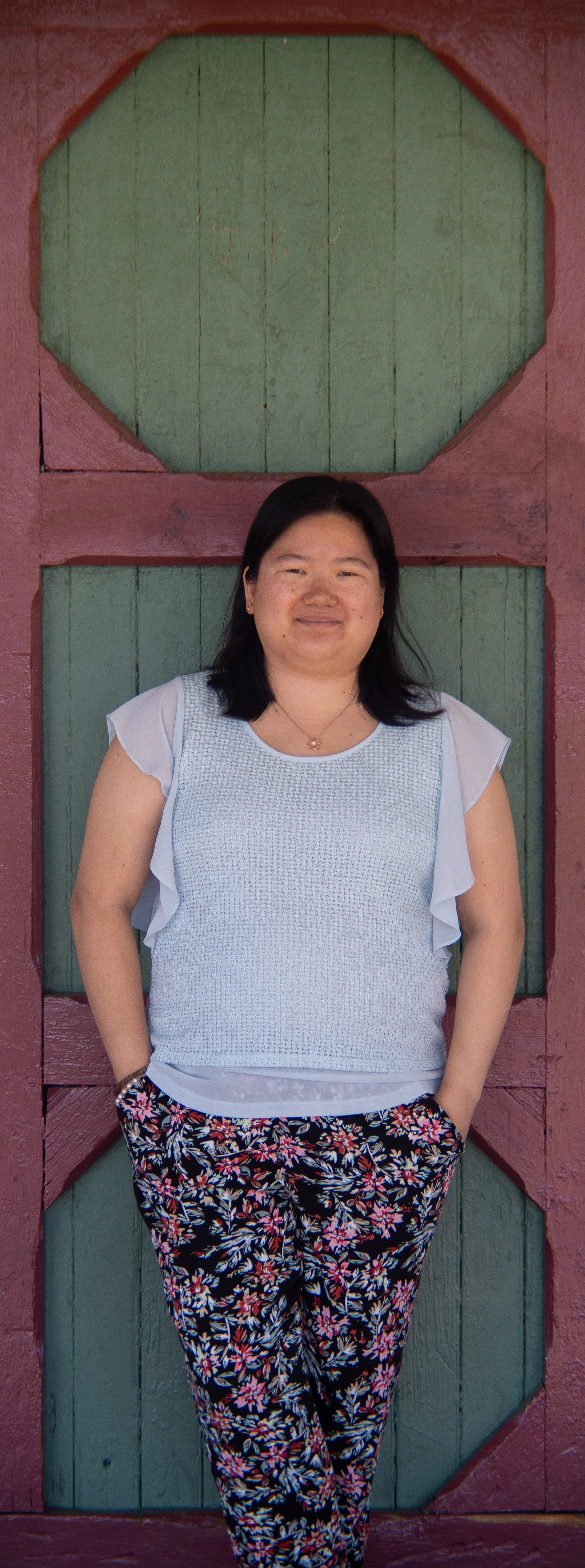 A woman standing in front of a teal and red wooden wall, smiling with her hands in her pockets, wearing a white top and floral pants.