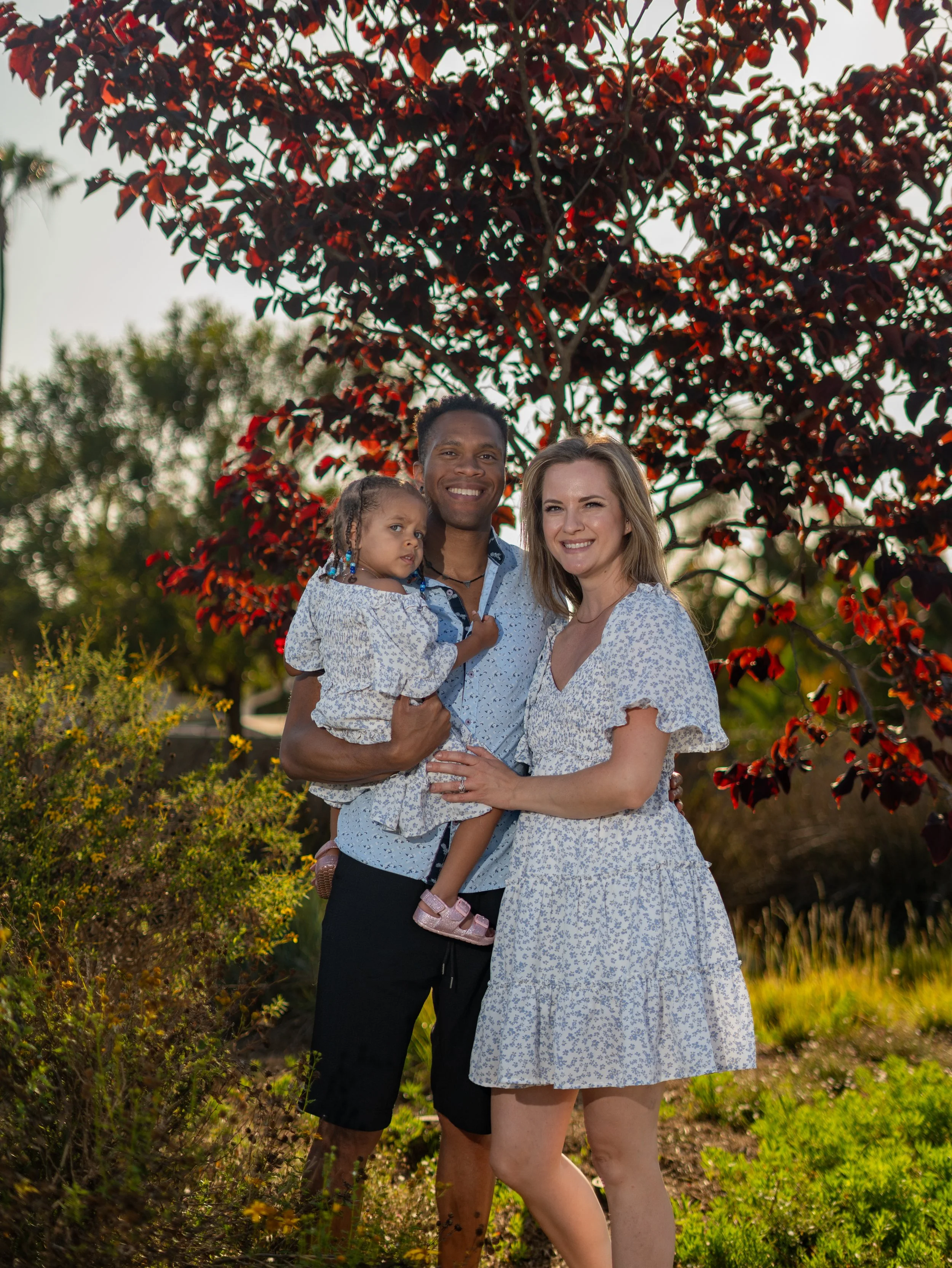 A happy family of three stands outdoors in front of red-leafed tree, with a man holding a young girl and a woman smiling at the camera. It is late afternoon or early evening sunlight.