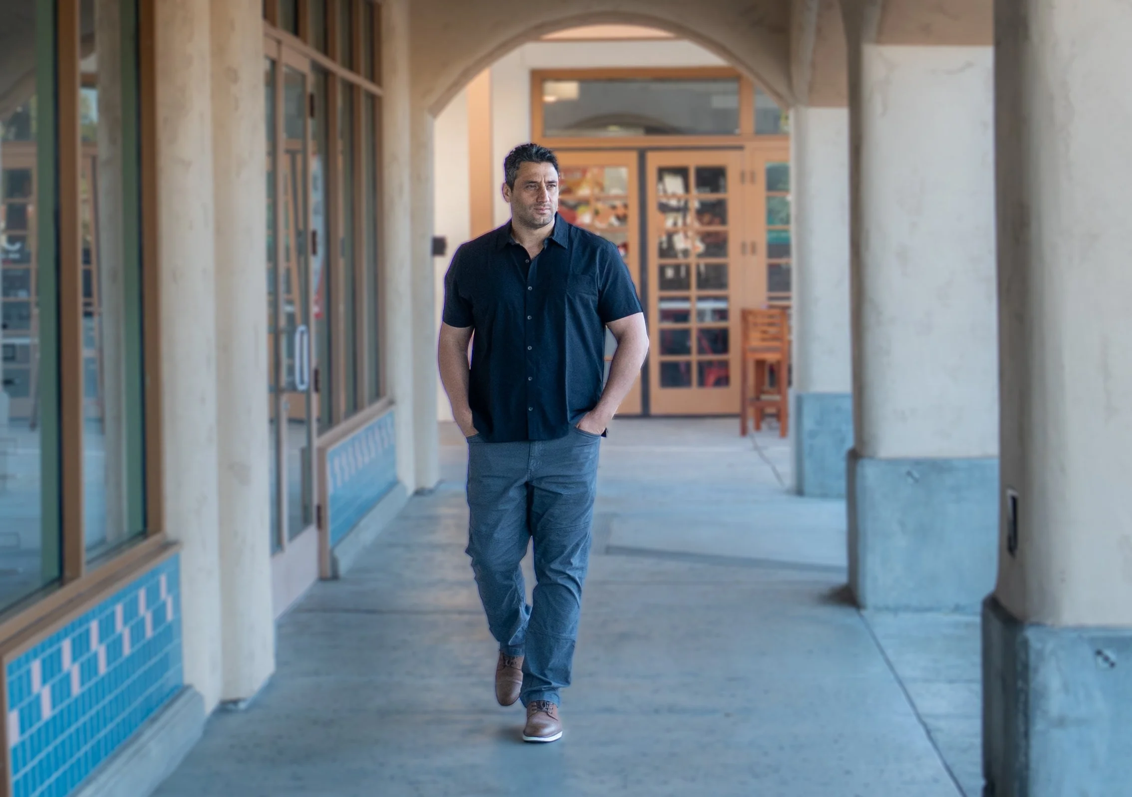 A man walking down a covered outdoor walkway with his hands in his pockets, wearing a black shirt and gray pants.