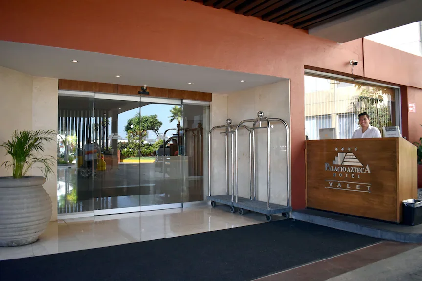 Hotel reception desk with a staff member behind it, luggage carts, and a large potted plant near glass sliding entrance doors, with outdoor greenery visible through the doors.
