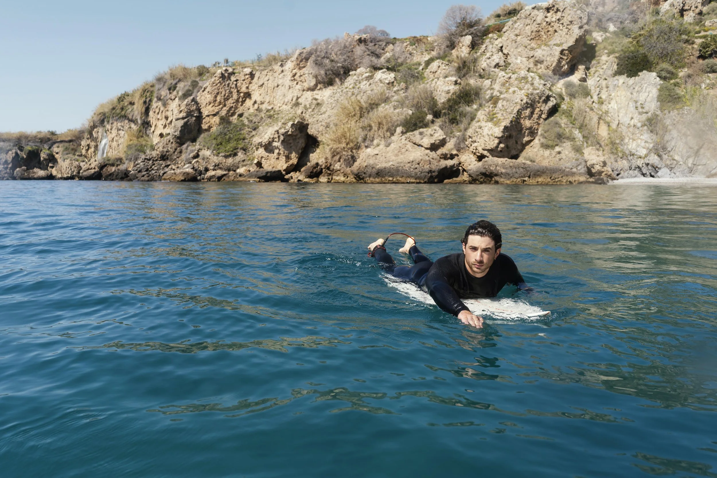 A man lying on a surfboard in the water near rocky cliffs with sparse vegetation, during daylight.