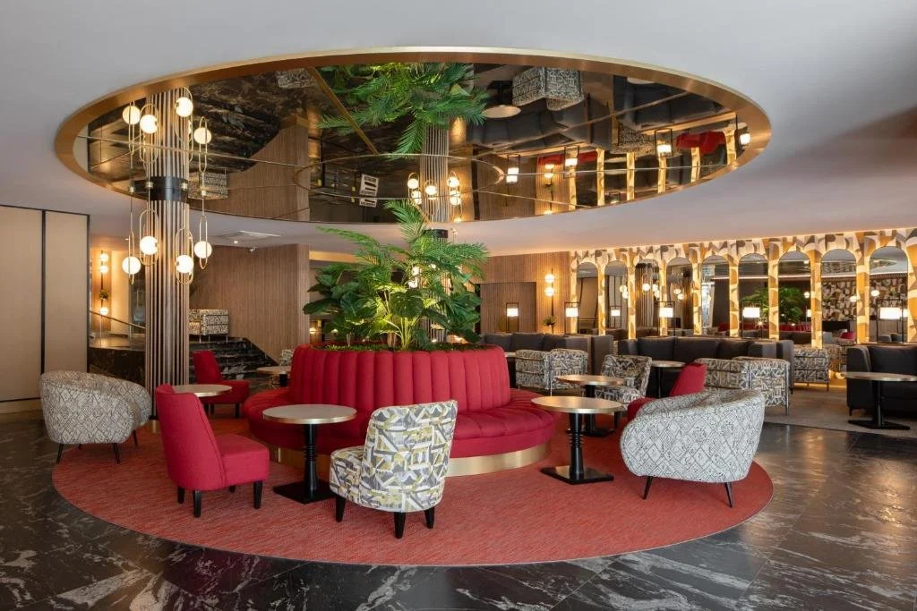 Elegant hotel lobby with a circular seating area, central greenery, and modern lighting fixtures, featuring a mix of red and patterned chairs.