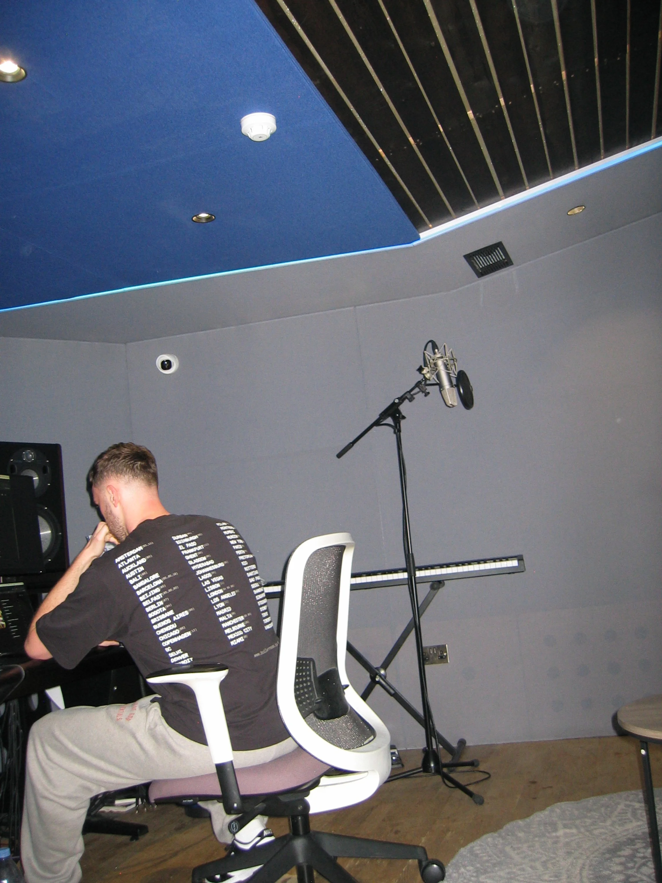 A person is sitting on an office chair in a recording studio, with a microphone on a stand, a keyboard, and speakers in the background. The ceiling has a painted design with blue, black, and gray sections.