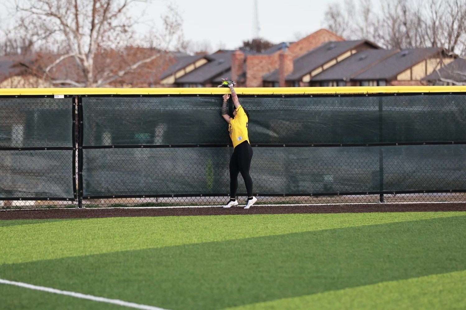 A person in sports attire hanging onto a green fence beside a grassy field, with residential houses and leafless trees in the background.