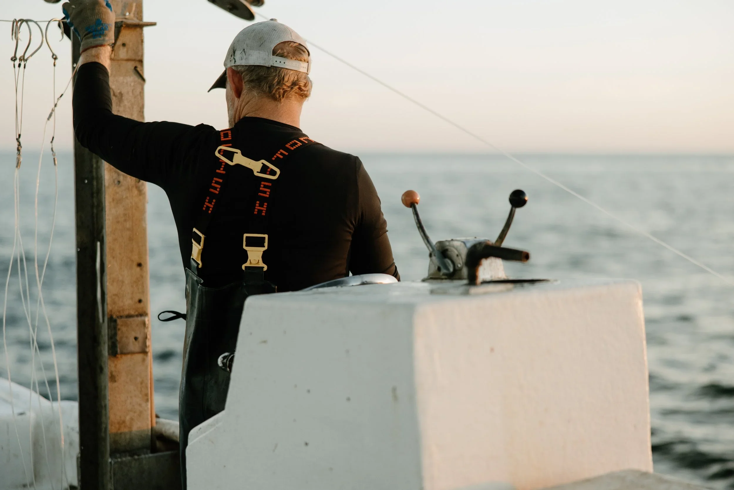 A man wearing a backwards cap and black shirt appears to be fishing on a boat in the ocean during sunset.