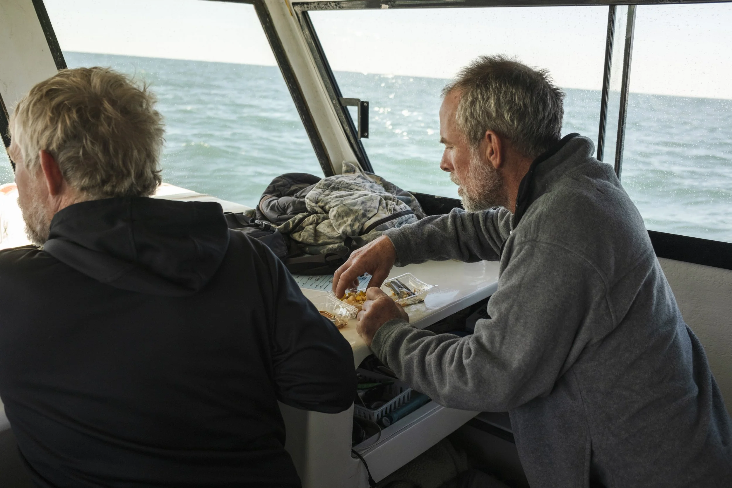 Two men with gray hair and beards sitting inside a boat cabin, looking out at the ocean through the windows. One man is eating snacks from a plastic container on the table.