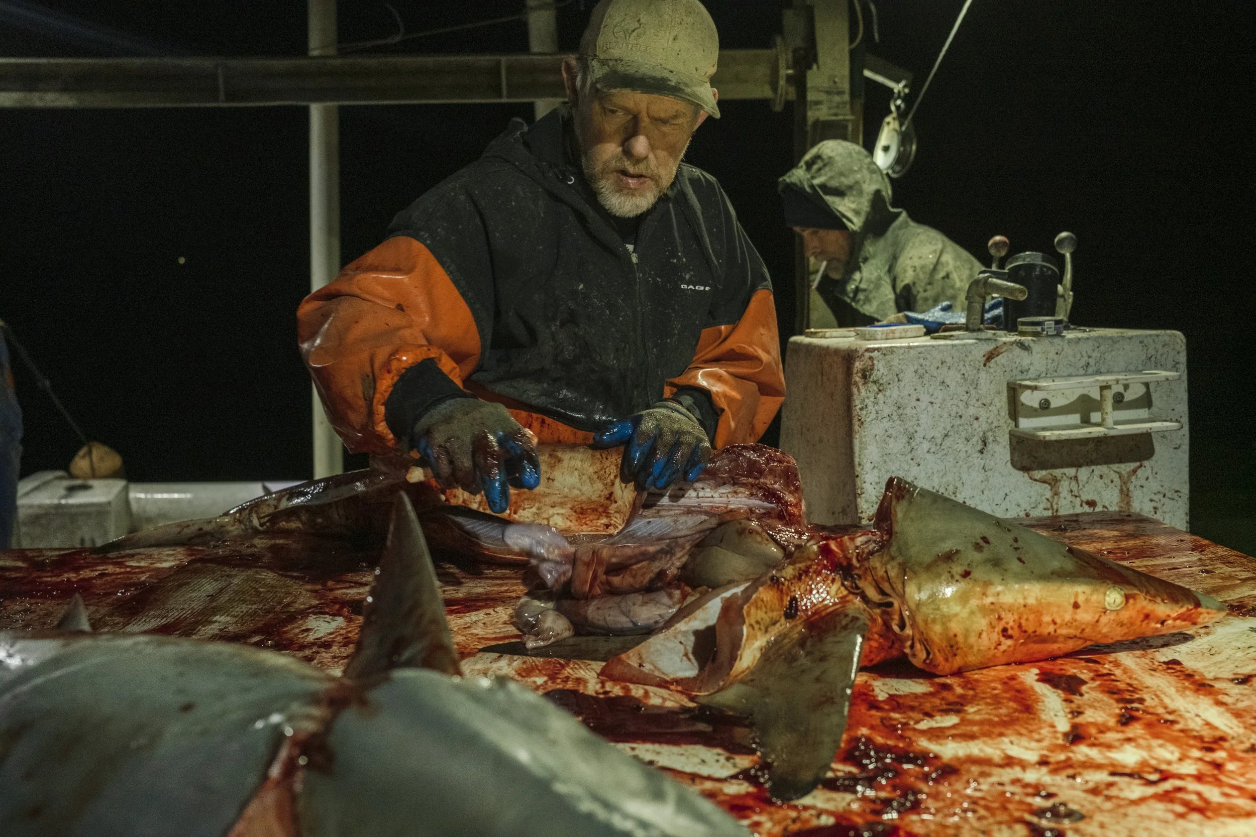 Two fishermen are cleaning a large fish on a wet table outdoors at night, with blood and fish parts scattered around.