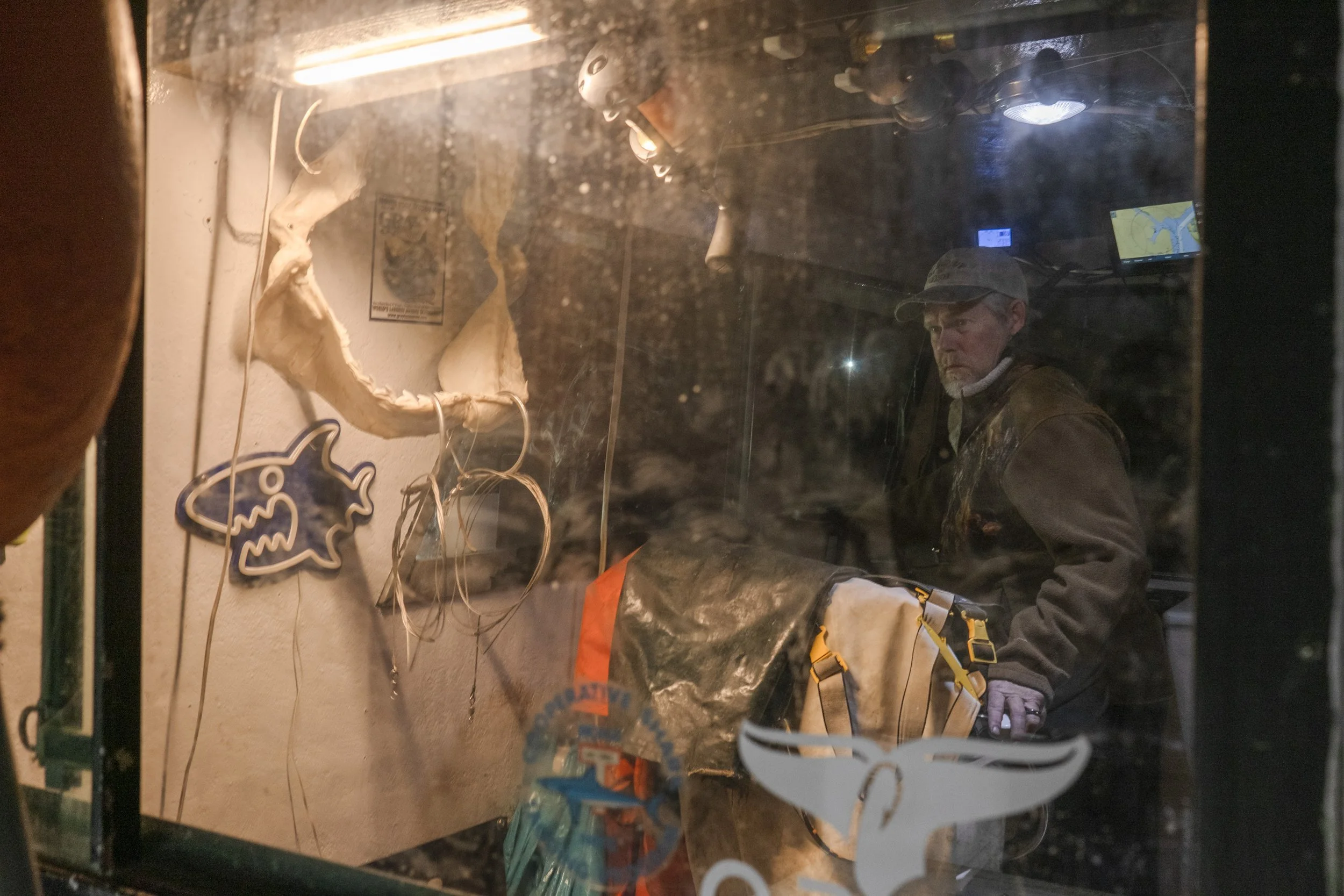A man wearing a camouflage cap and brown jacket is seen through a shop window, with various decorations and some illuminated screens inside.