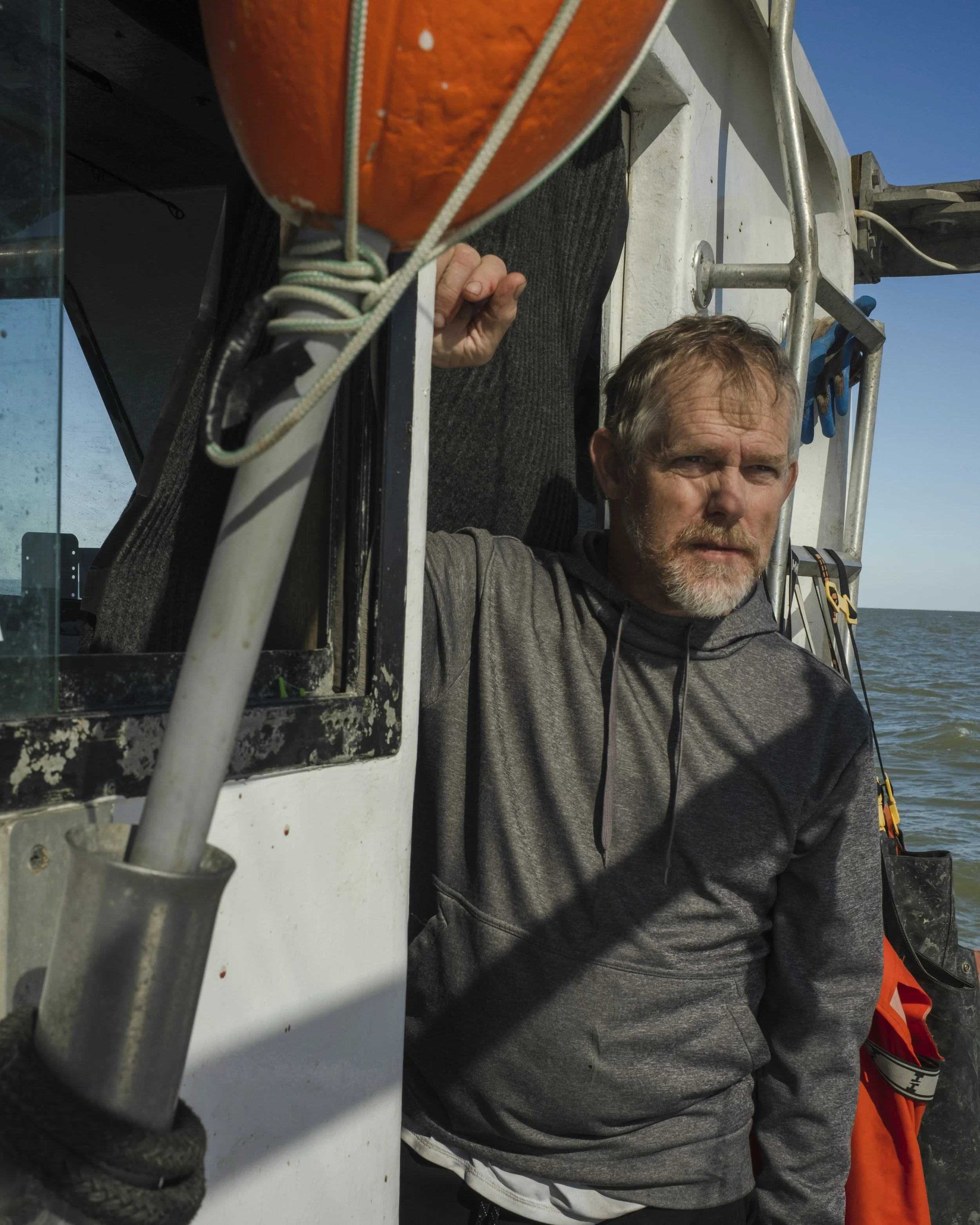 A man with a beard and grey hair standing on a boat, wearing a grey hoodie, seen against the ocean and blue sky.