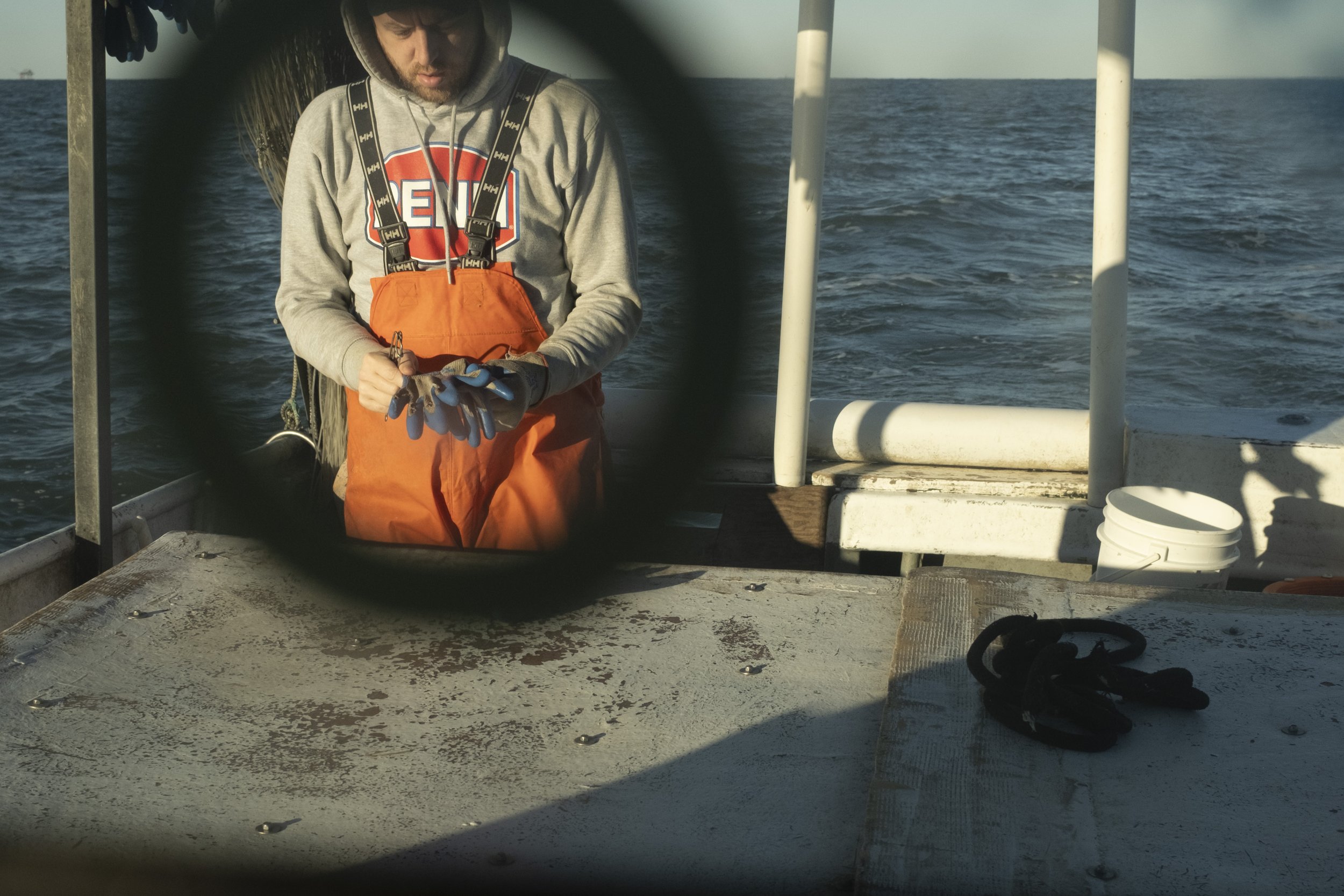 A man on a boat, wearing a gray hoodie and orange overalls, is working with gloves and tools. The photo is taken through a circular opening, with the ocean in the background and some equipment on the boat deck.
