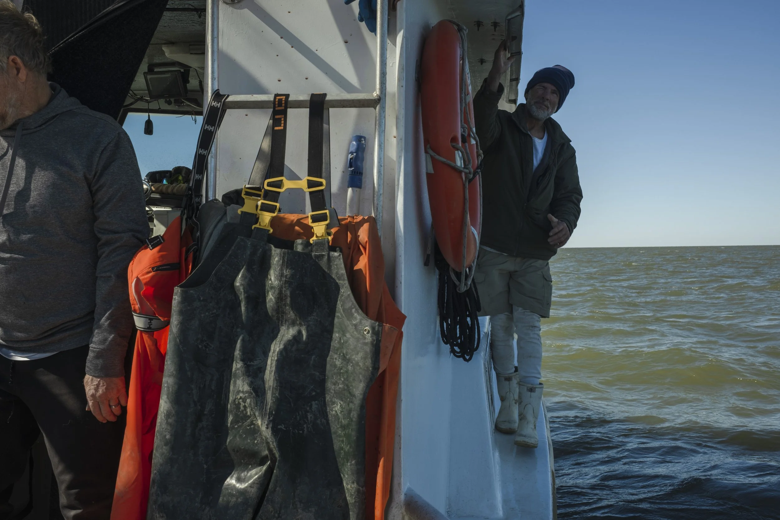 Two people are on a boat with water and sky in the background. One person is standing near the edge, smiling and waving, wearing a beanie, jacket, and shorts. The other person is partially in view, wearing a hoodie. The boat has safety equipment, inc
