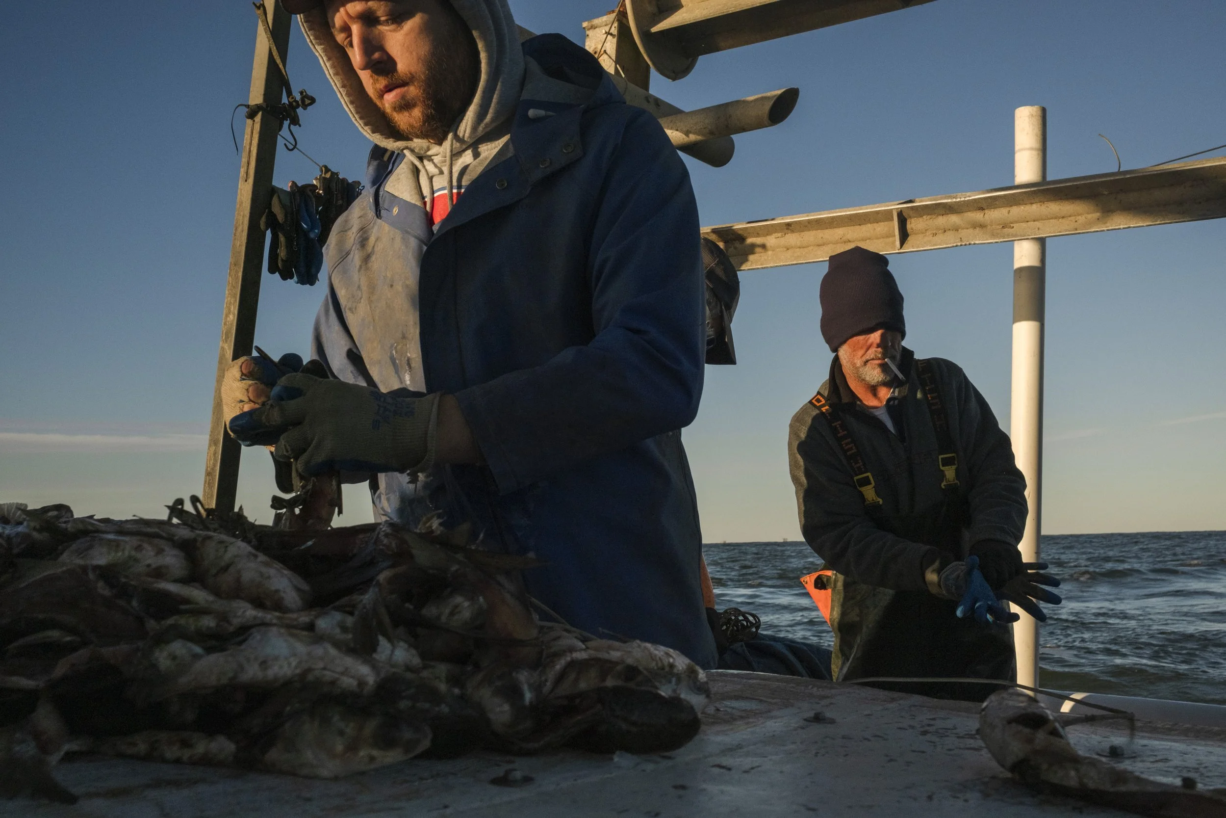 Two men engaging in fishing on a boat, with fish laid out on the surface. One man in a blue jacket is working with the fish, while the other man in a black beanie is standing nearby, smoking a cigarette. The scene is set at sea during sunset or sunri