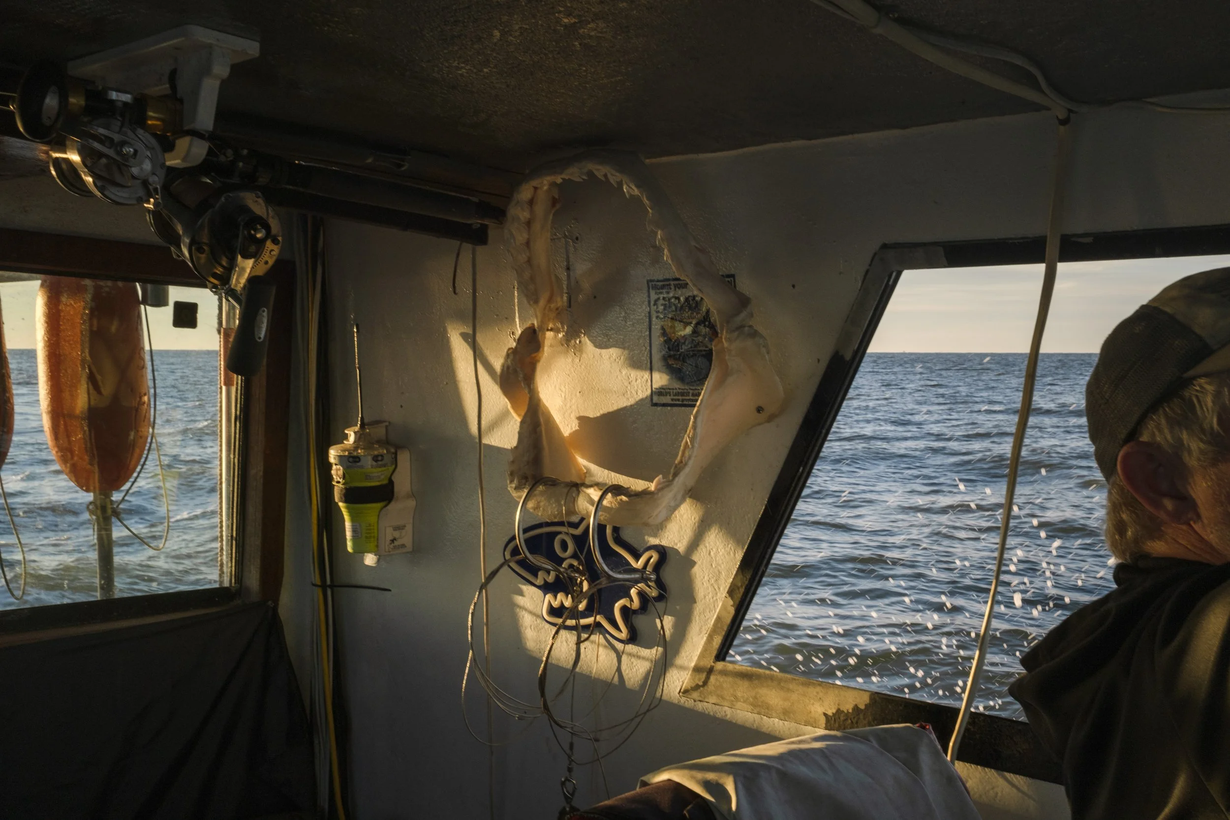 Interior of a boat with a view of the ocean through windows, showing fishing gear, life vests, and a man wearing a cap, near a large fish skeleton hanging on the wall during sunset.