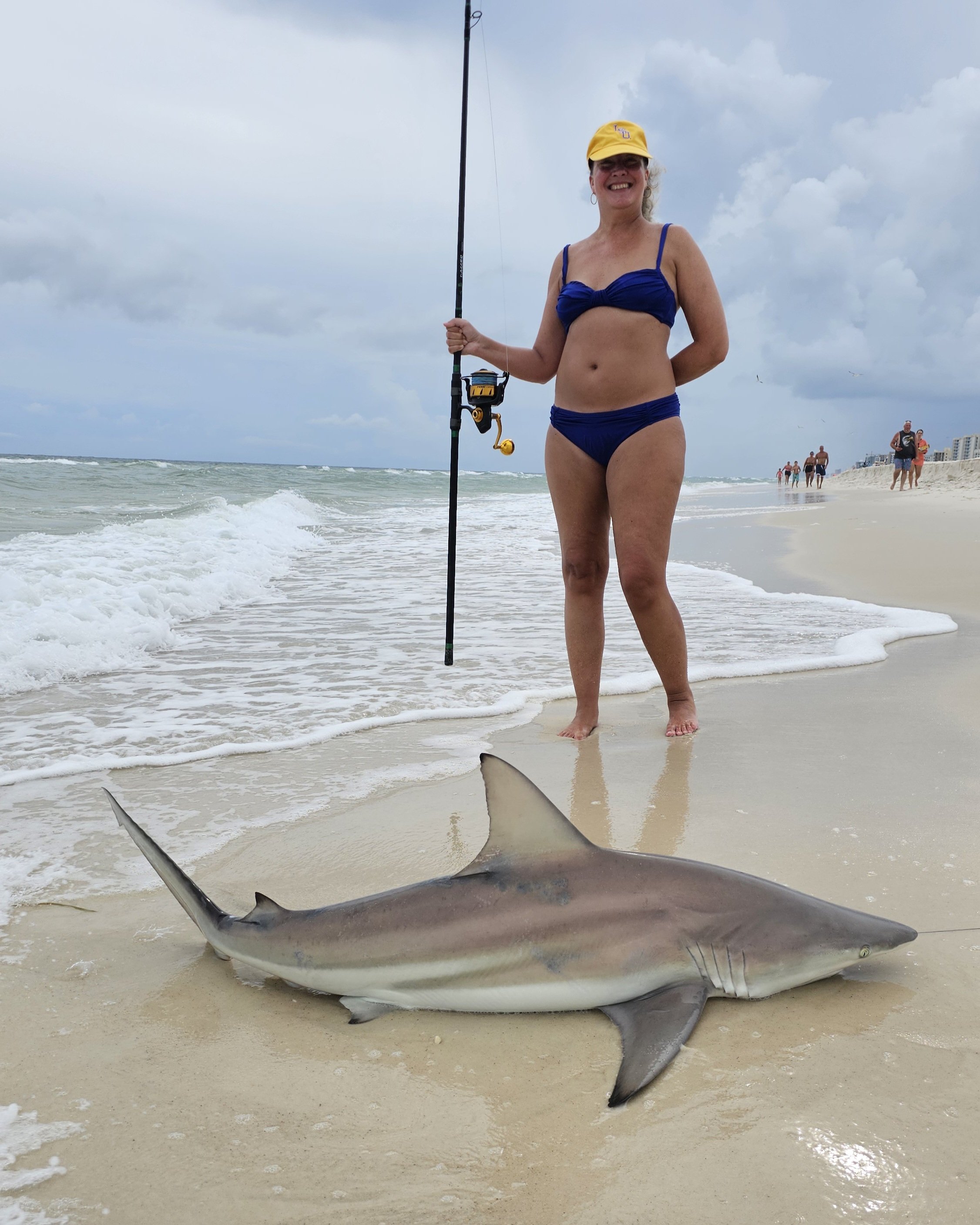 Woman in a blue bikini and yellow cap standing on ocean beach, holding fishing rod, next to a large shark on the sand, with cloudy sky and other beachgoers in the background.