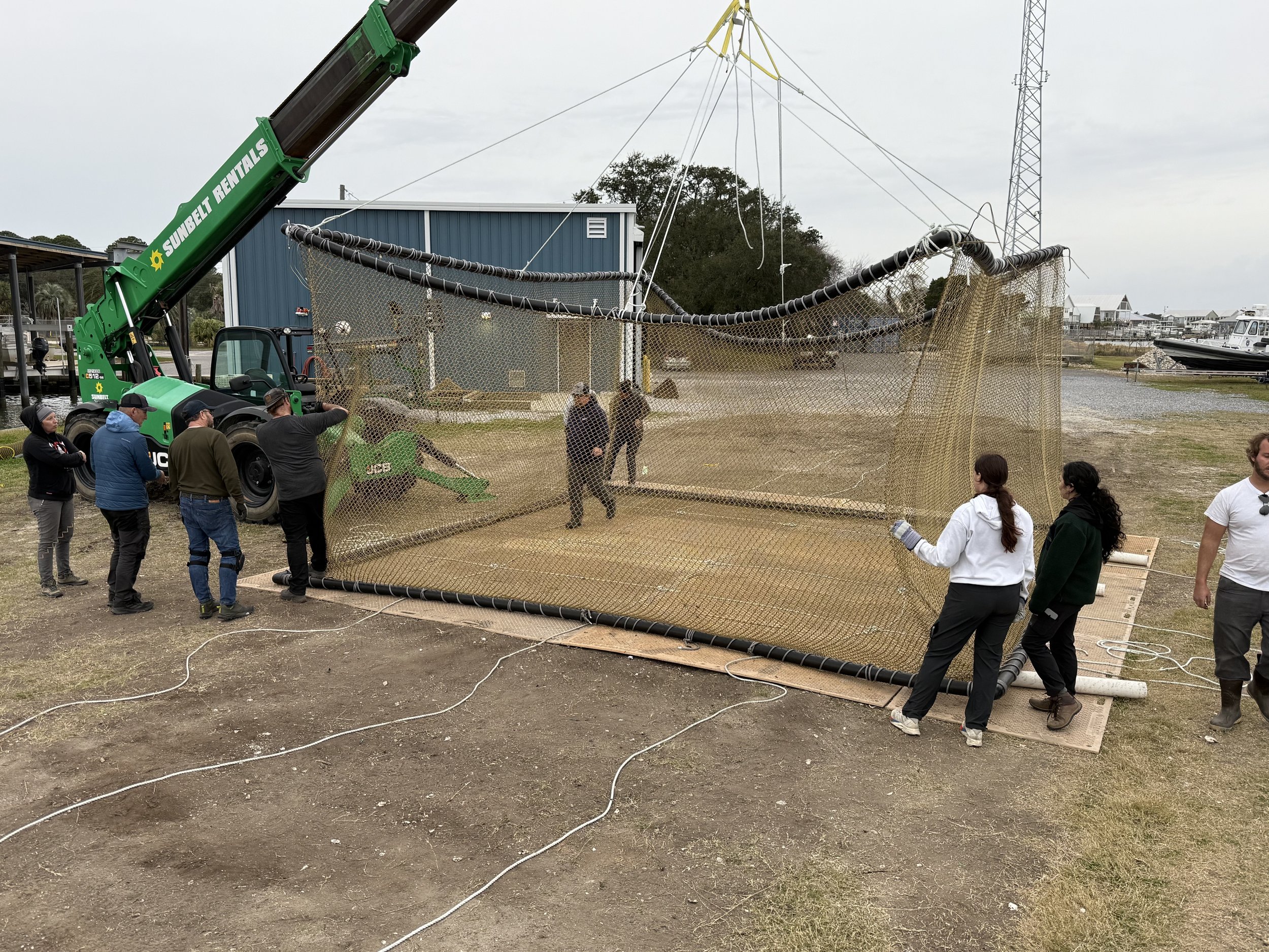 People assembling a large fishing net with a crane at a dockside area near boats and buildings.