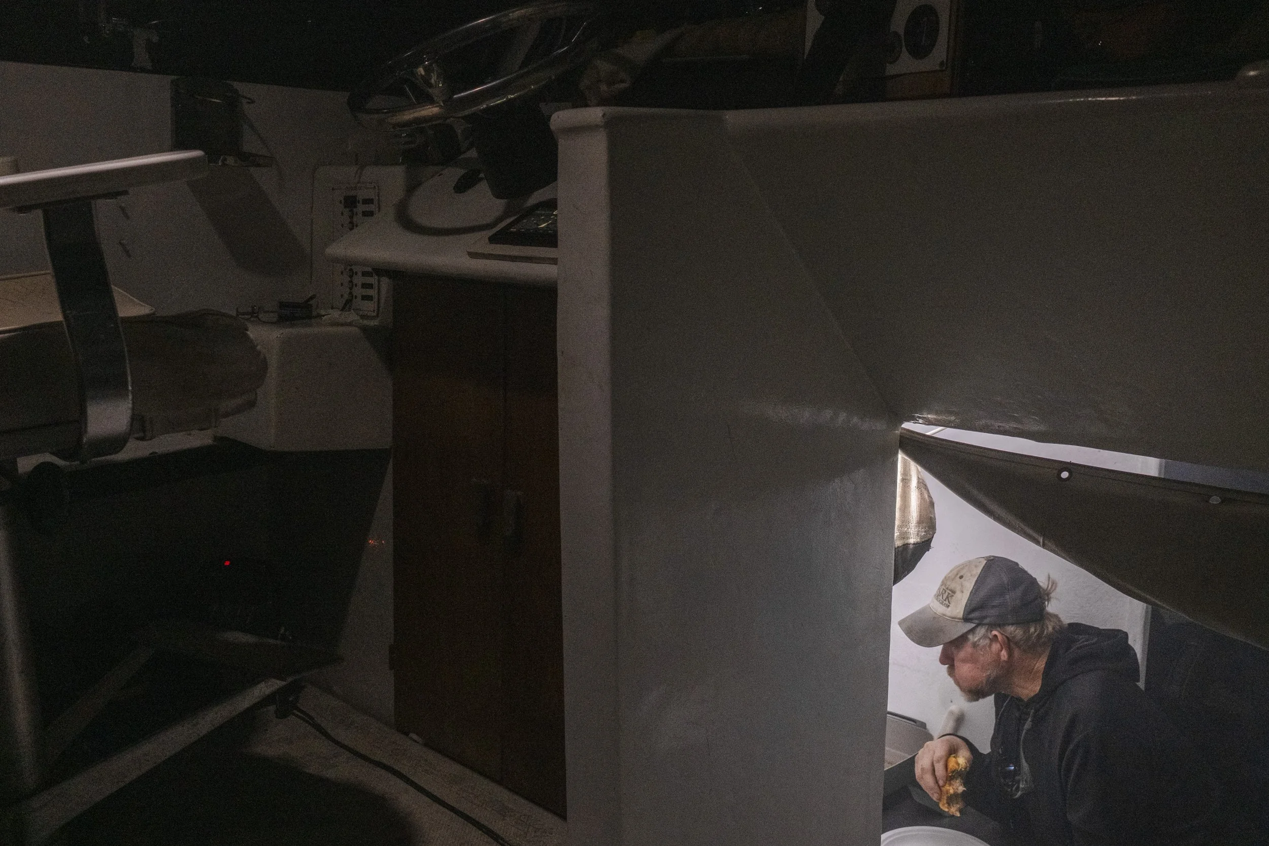 A man with gray hair, glasses, a tan baseball cap and black hoodie sitting on the floor next to a toilet, holding a piece of pizza and looking down.