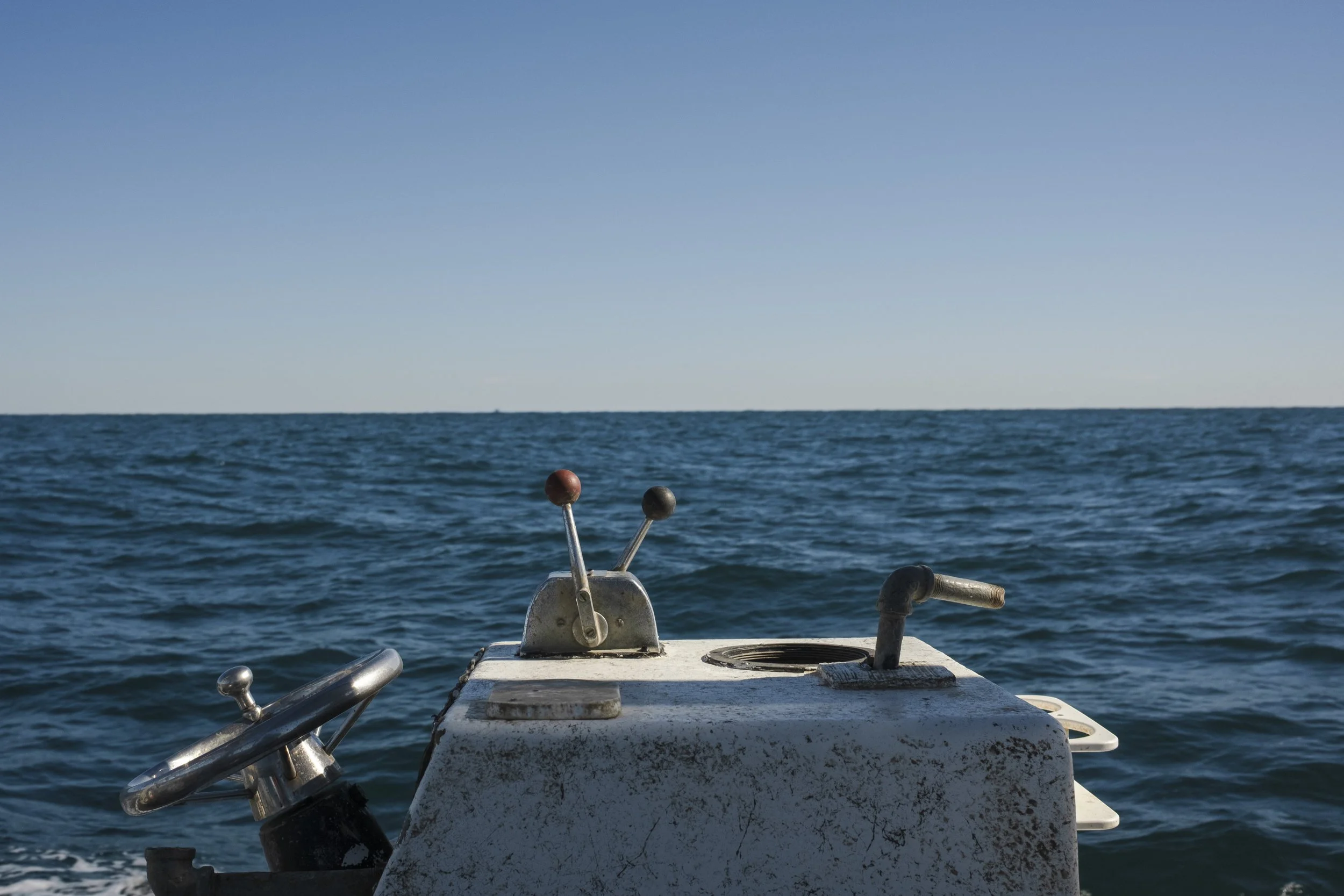 View from a boat showing a steering wheel and control levers with an ocean and clear blue sky in the background.