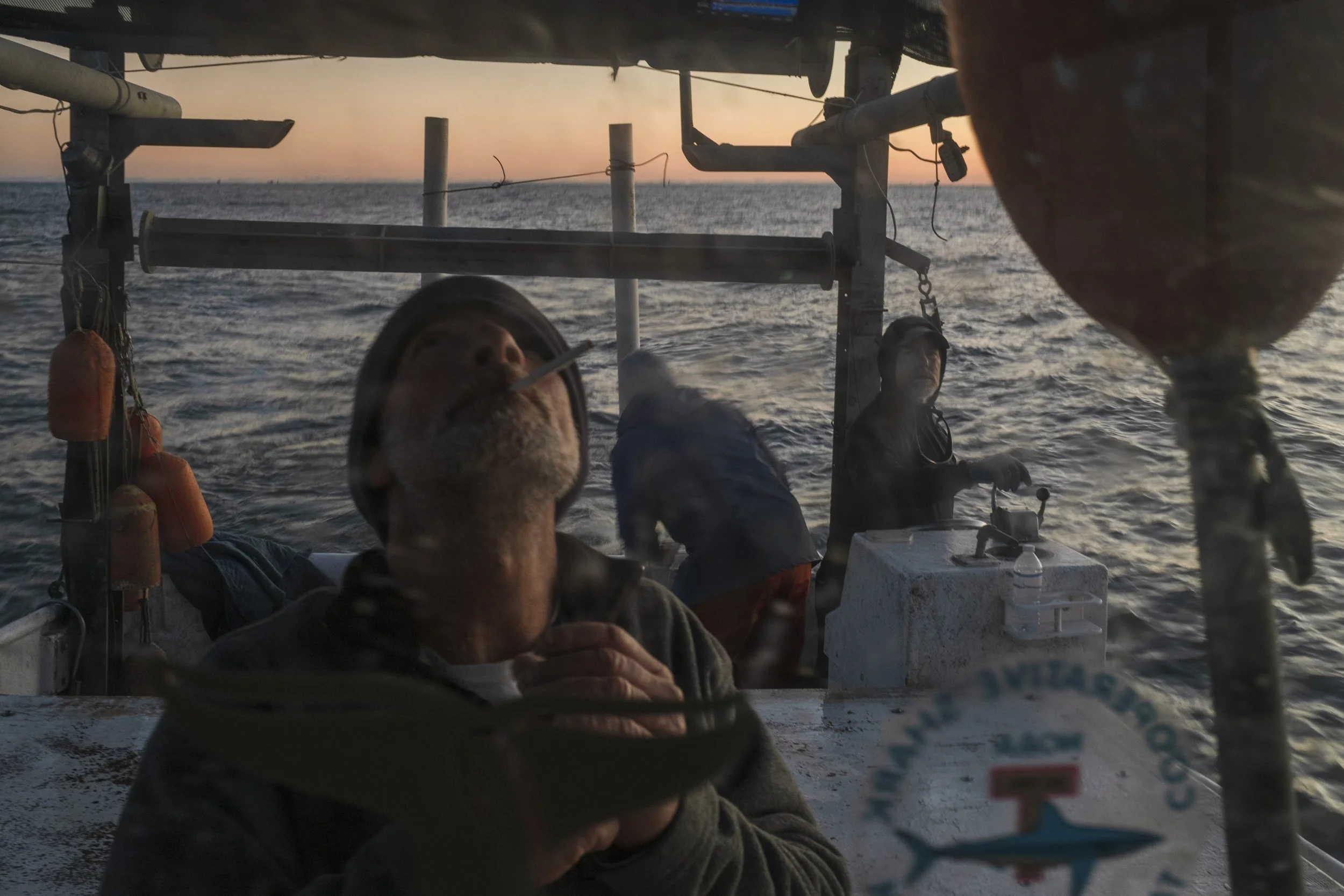 Three men on a boat at sea during sunset. One man is smoking a cigarette and wearing a hat, while another is looking at the camera. The third man is in the background by the boat controls. The water is calm, and the sky is a mix of orange and blue hu