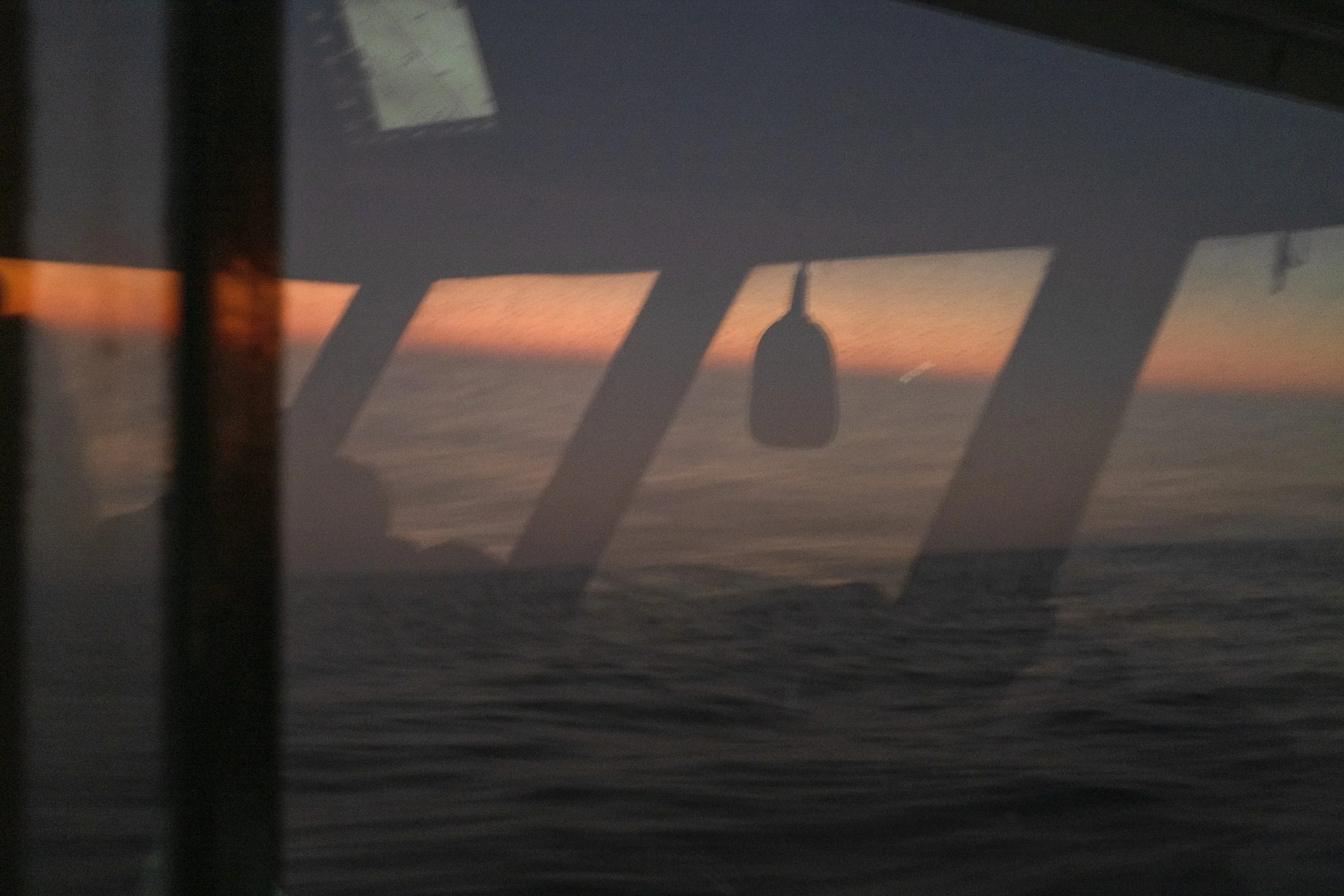 View of a sunset over the ocean through an aircraft window with reflections and a hanging nose of the aircraft.