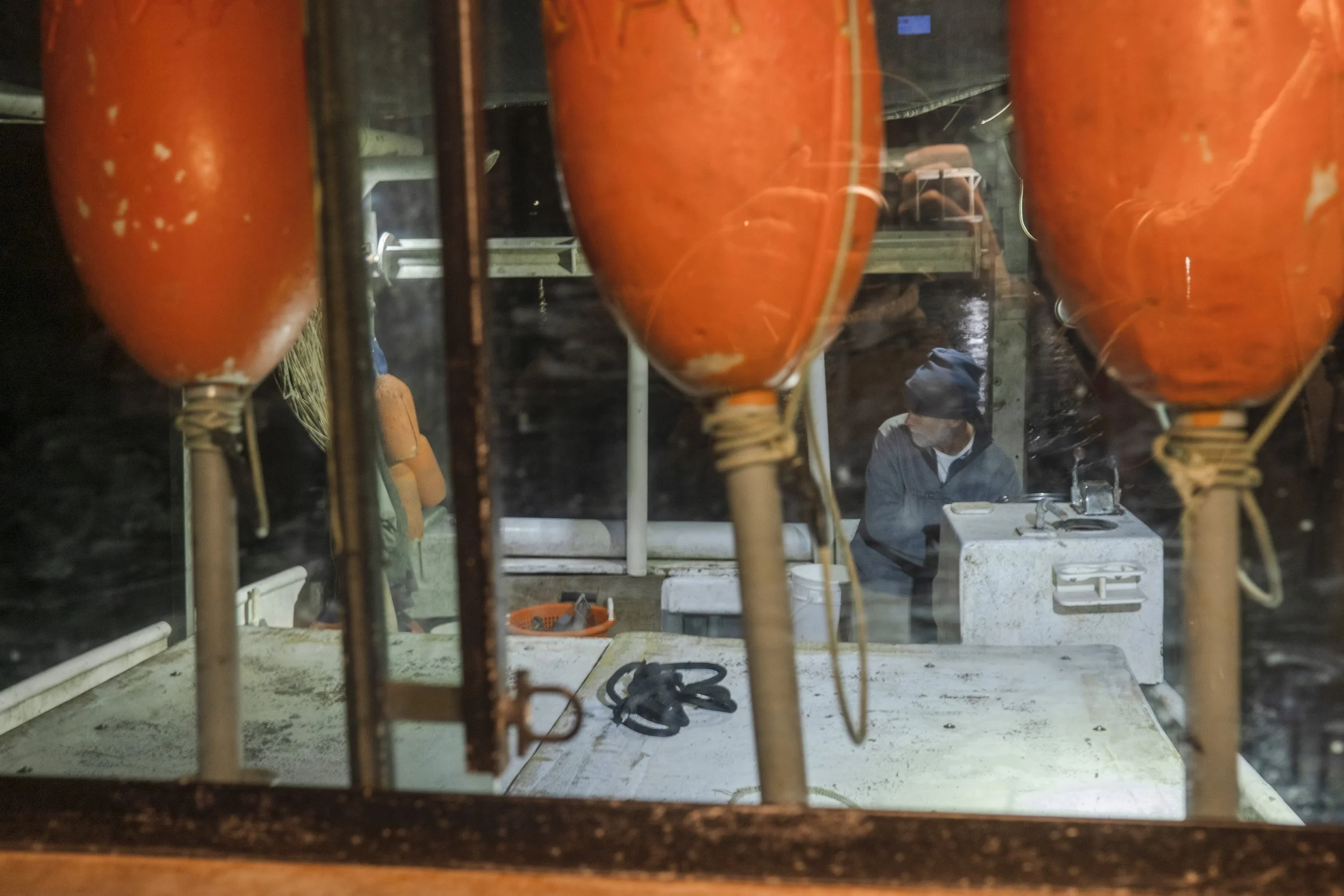 A view through a window with orange buoys hanging, showing two fishermen or workers on a boat, one wearing a dark jacket and a head covering, the other with an orange jacket, working or talking near a white cooler or storage box on deck.