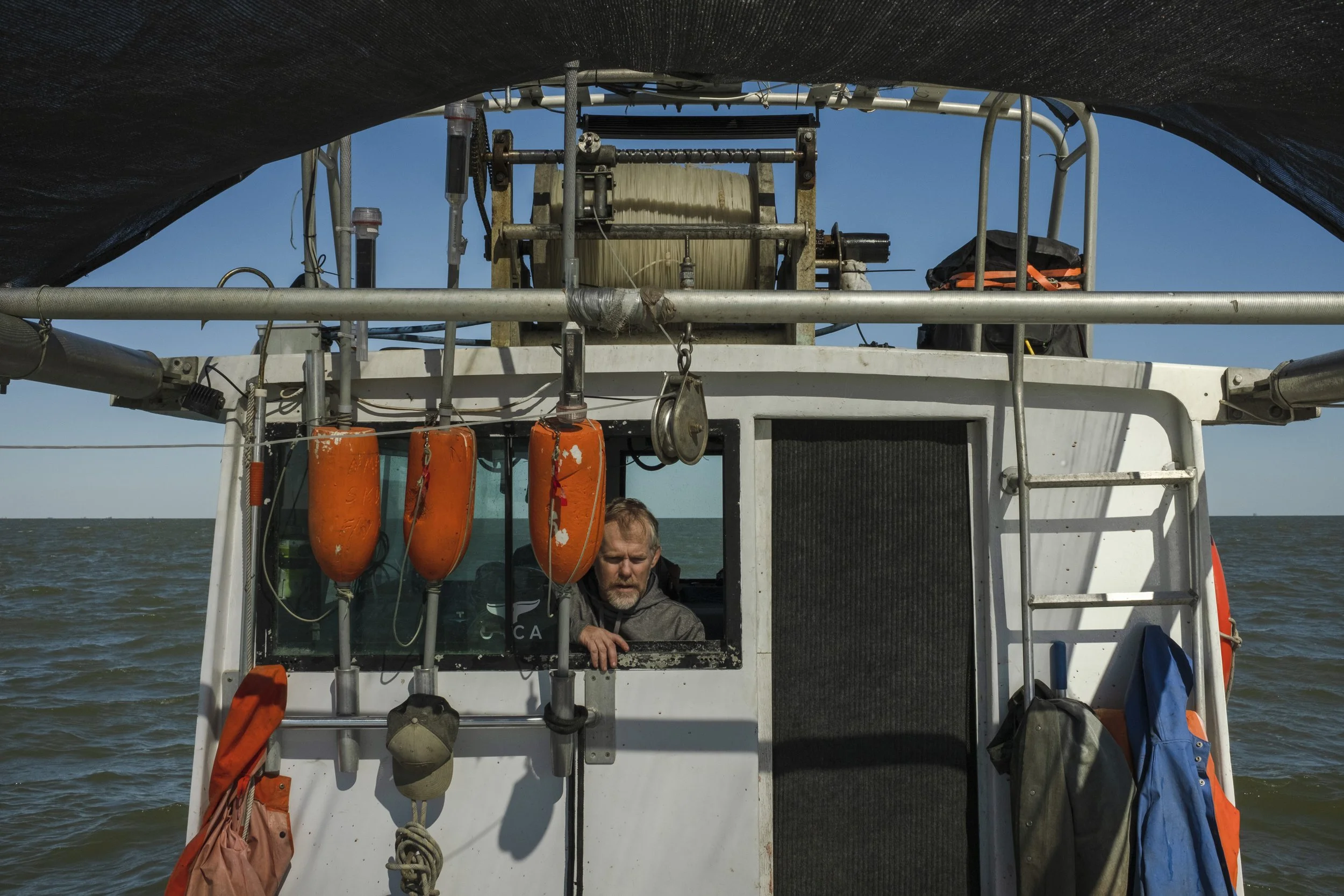 A man looking out of the window of a boat, with sea and clear sky in the background. The boat has orange buoys hanging on the front, and various ropes and equipment on top.