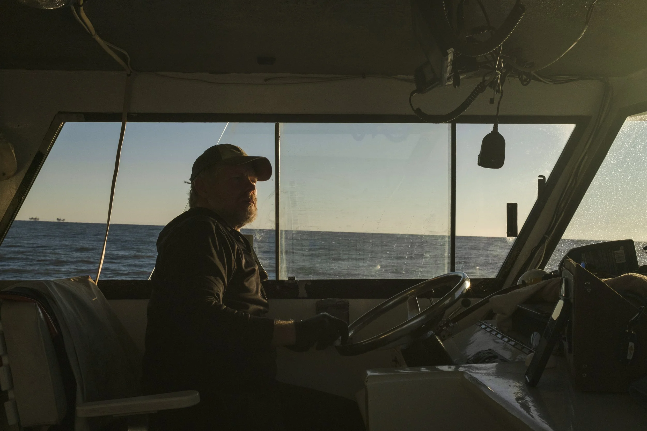 Man with a beard, wearing a cap and dark clothing, sitting in the cockpit of a boat, steering through the water at sunset.